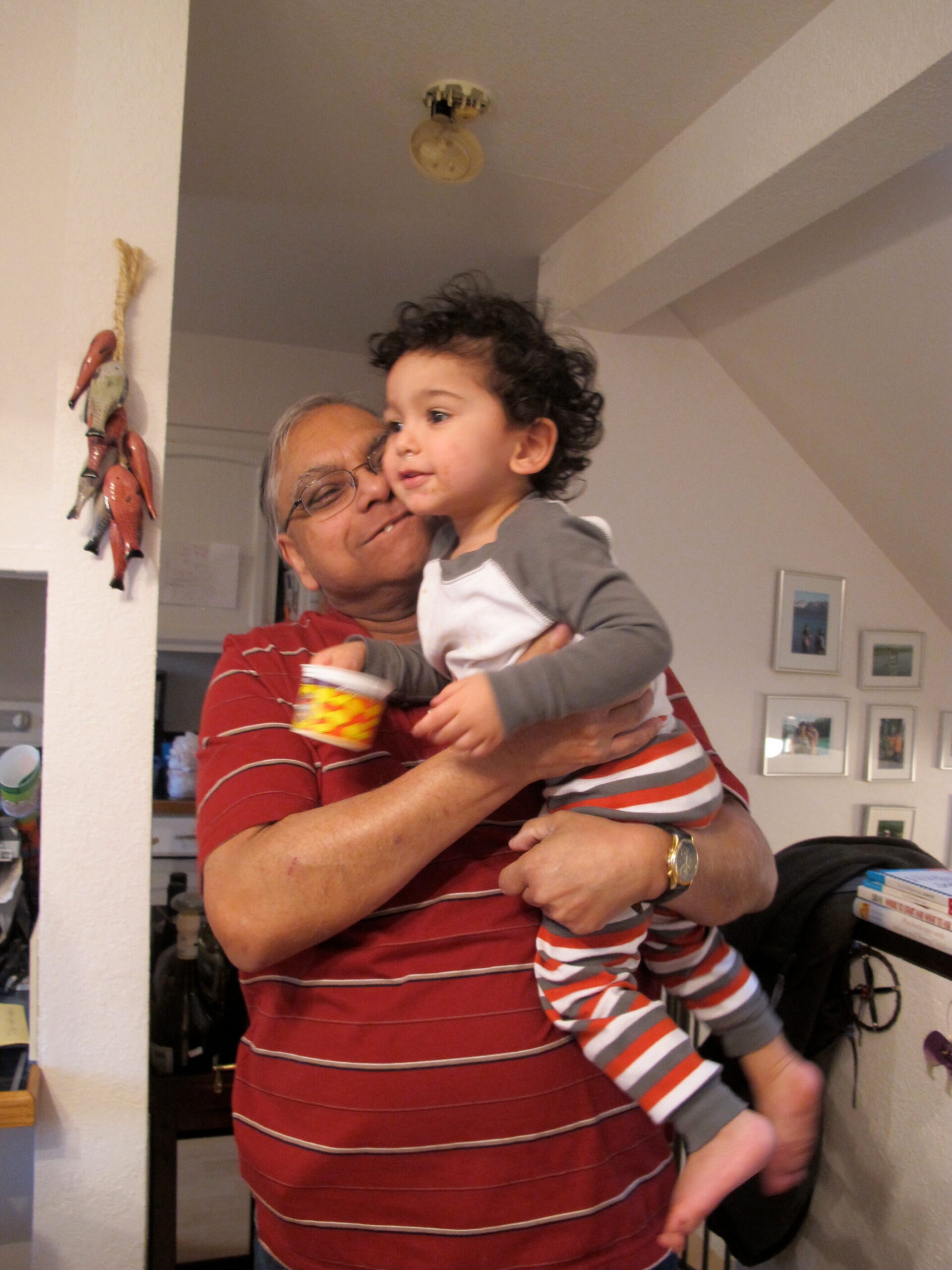 Grandfather wearing a red striped shirt holds a smiling toddler with curly hair indoors in a kitchen area.