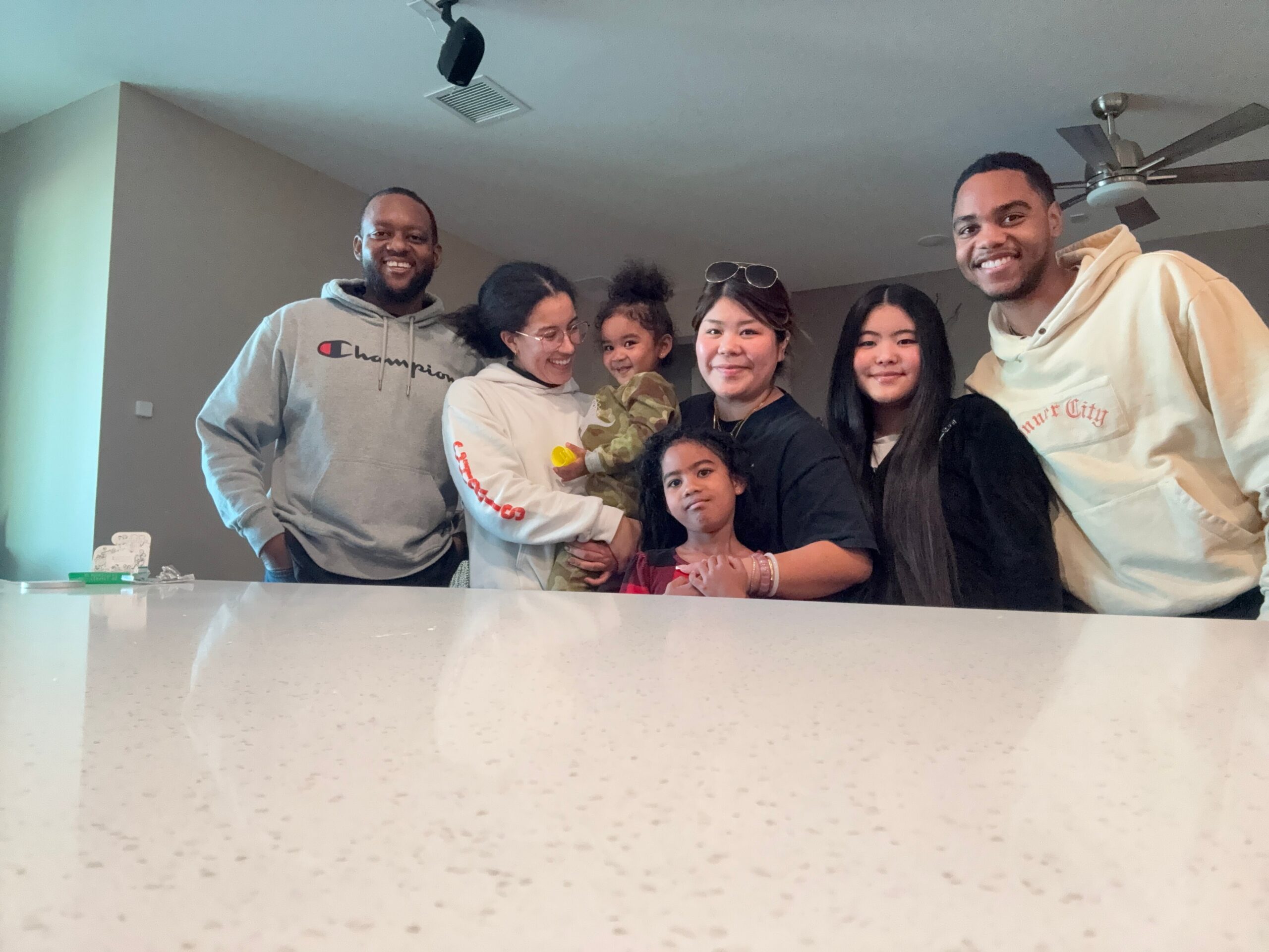 A group of smiling adults and children posing together indoors, standing near a kitchen counter under a ceiling fan.
