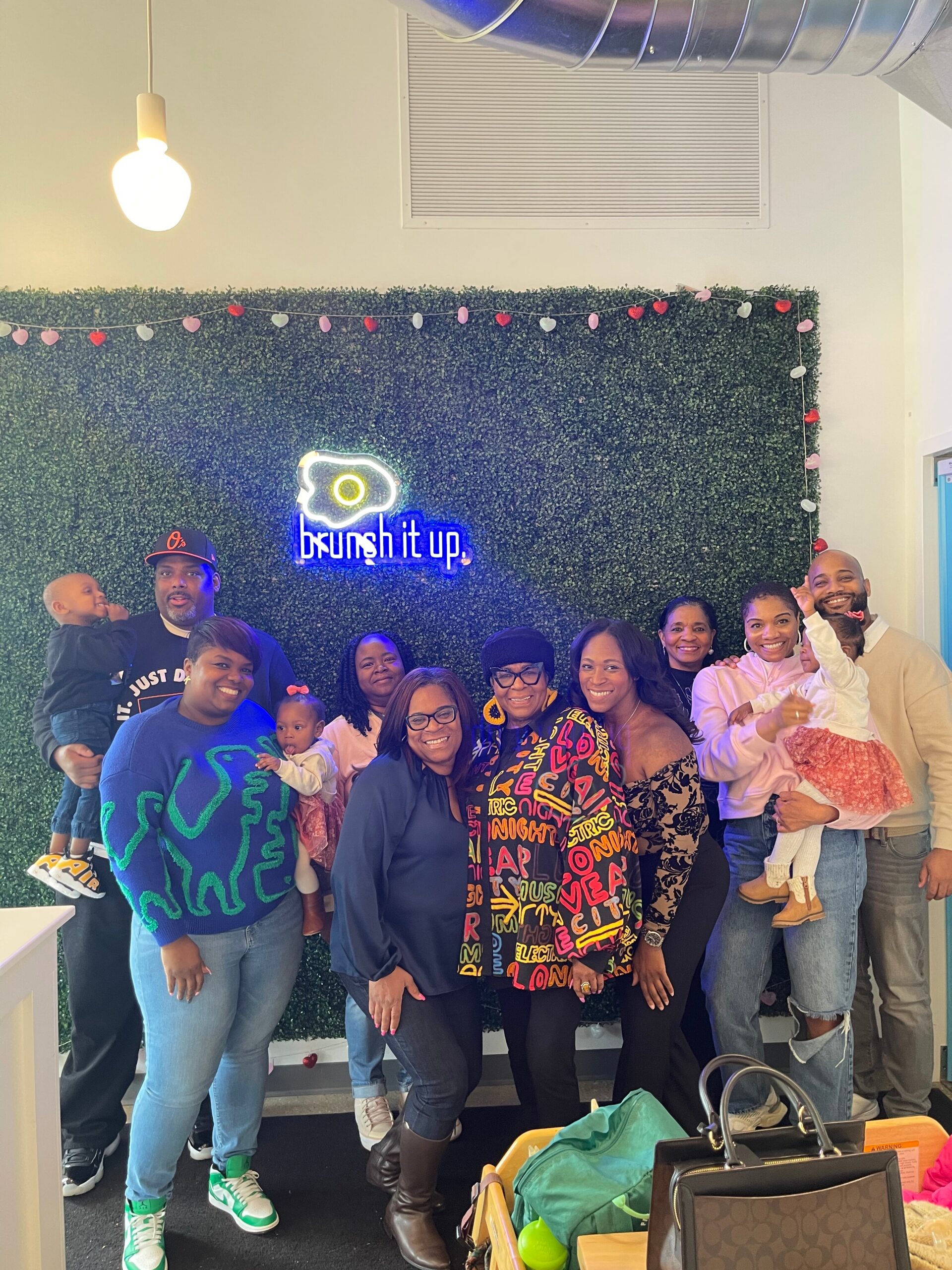 Group of diverse adults and children posing for a photo in front of a green hedge backdrop with a neon 'brush it up' sign and heart garland.