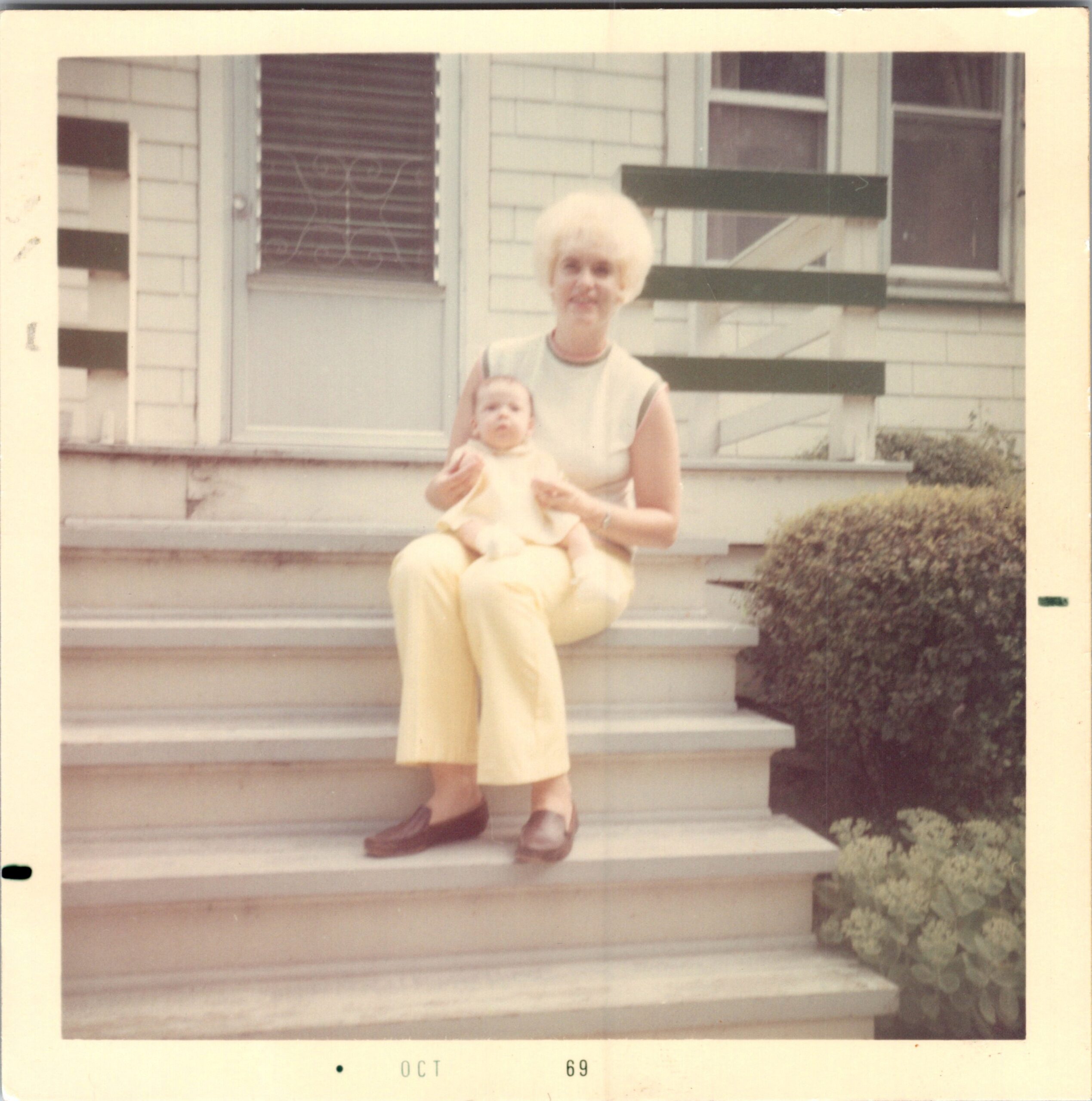 Woman with short blonde hair sits on front steps holding a baby outdoors in a light-colored dress, in front of a house with windows and shrubbery.