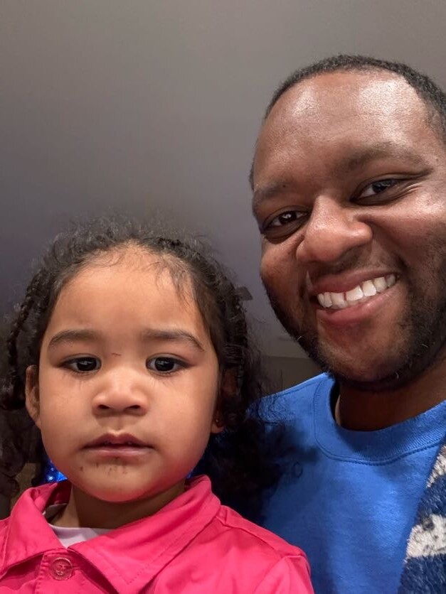 Smiling man in a blue shirt taking a selfie with a young girl in a pink jacket, indoors.