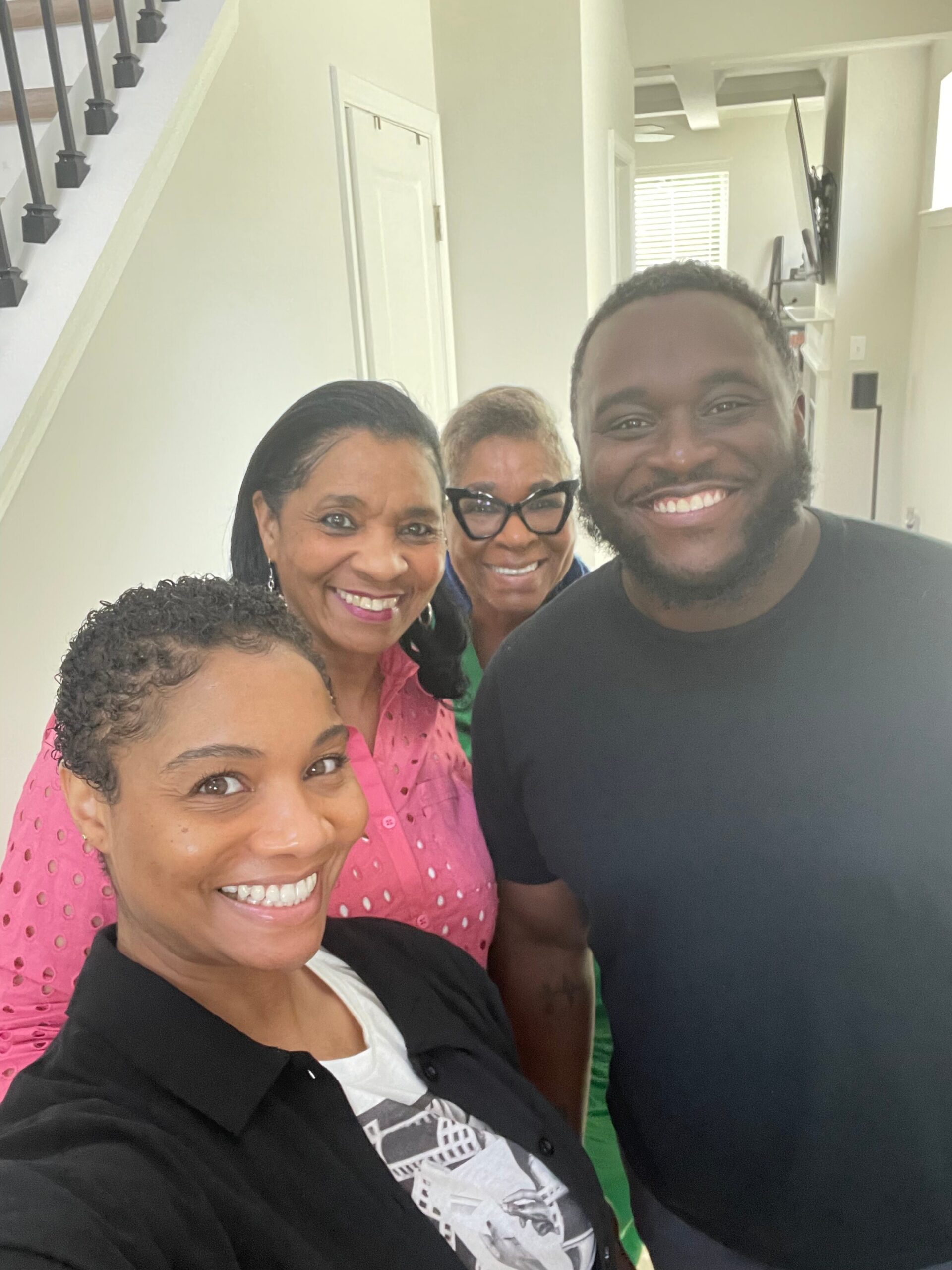 Four people smiling and posing for a selfie in a bright hallway near a staircase.