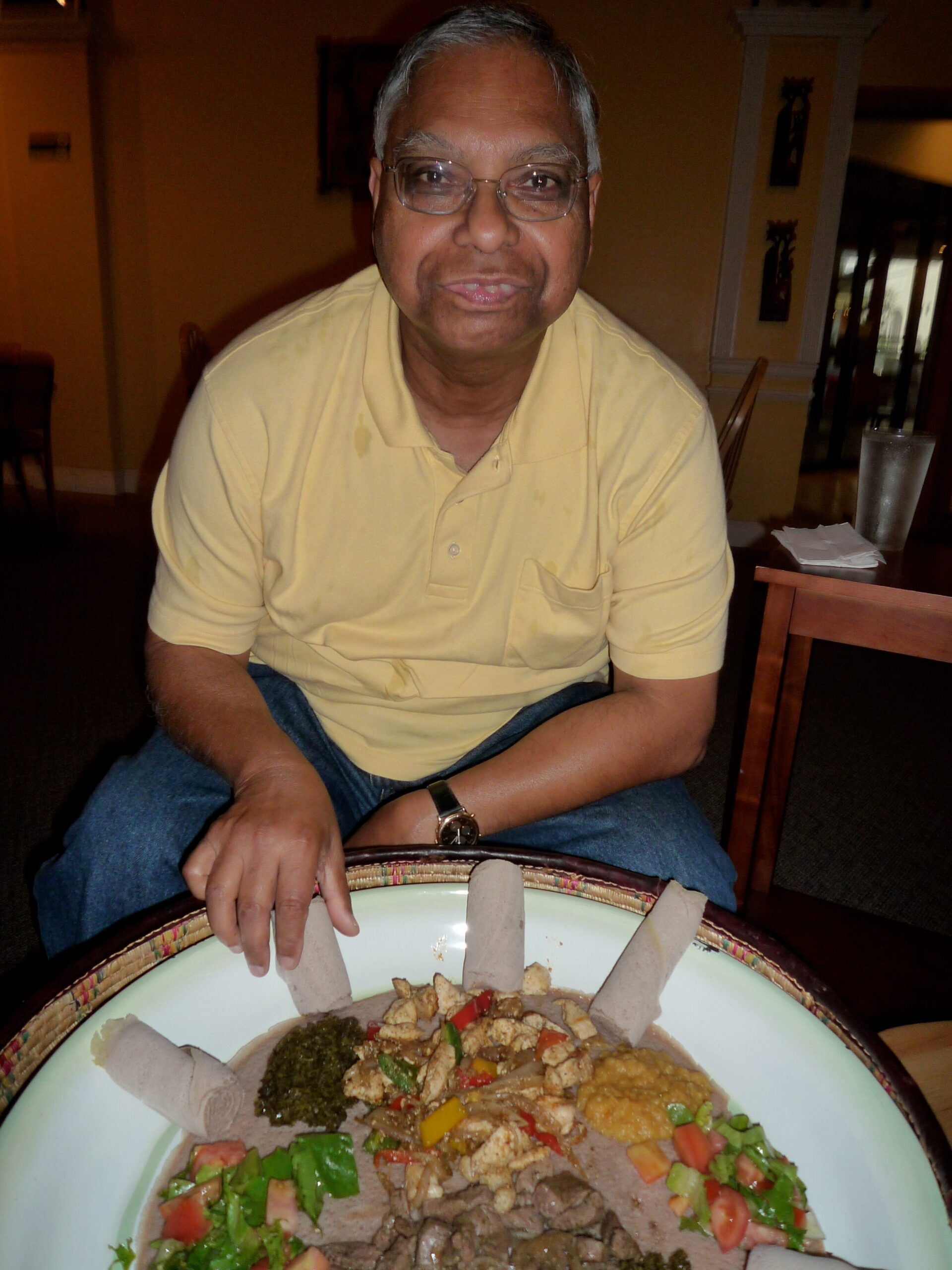 Man wearing glasses and a yellow polo sits at a table with a large plate of mixed Mexican food in front of him.