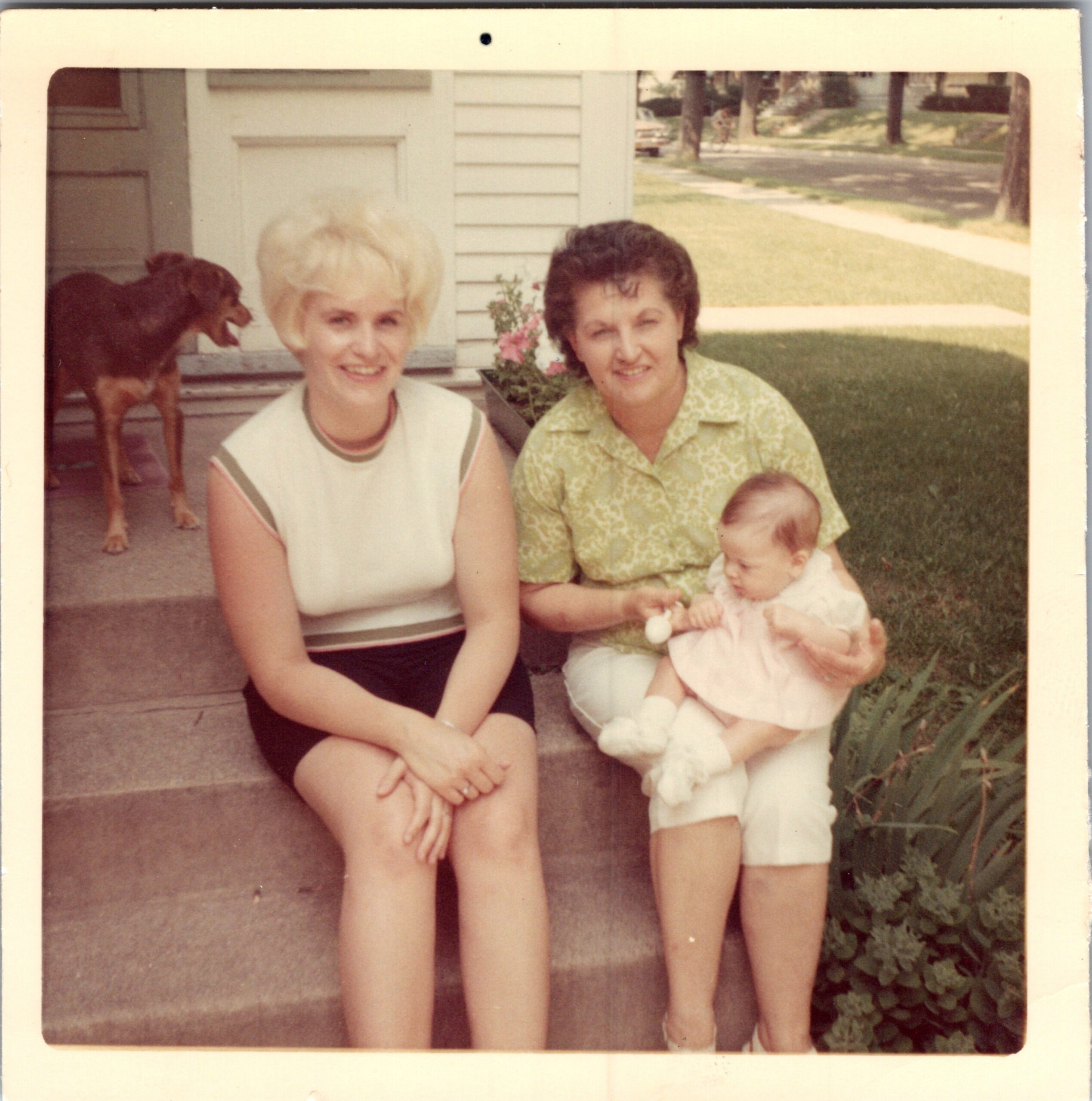 Two women pose on a front porch with a baby, a brown dog standing on the left in the background.