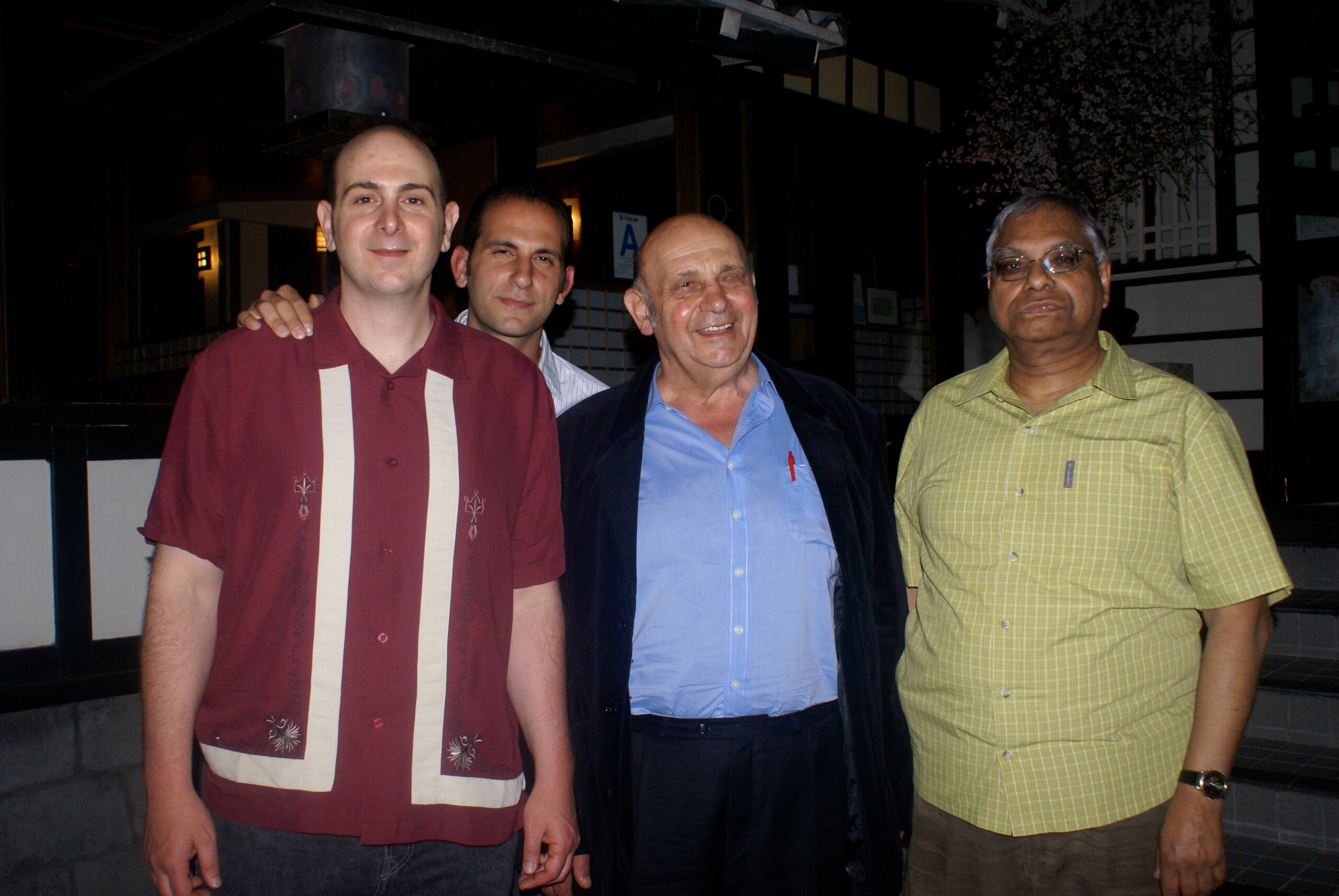 Four men pose for a photo indoors at night, smiling at the camera with arms around each other behind the first man on the left.