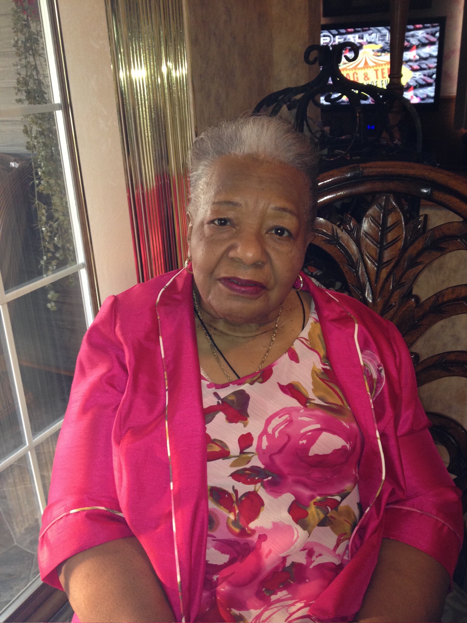 Smiling elderly woman with short gray hair in a pink jacket and floral dress, seated near a glass door.