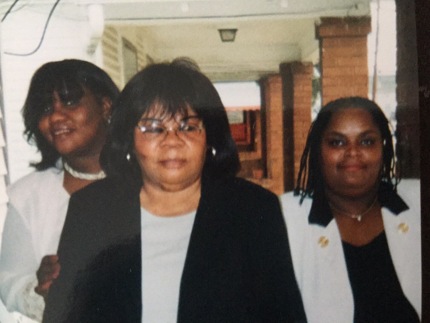 Three women stand together in a hallway, wearing white tops with black jackets; the middle woman wears glasses.