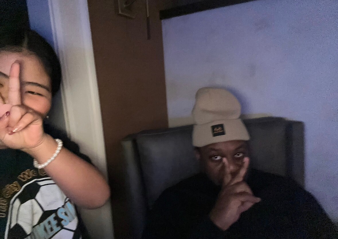 Two friends posing in a dim room: one shows a peace sign near their face, the other wears a beige hat and gestures for silence.