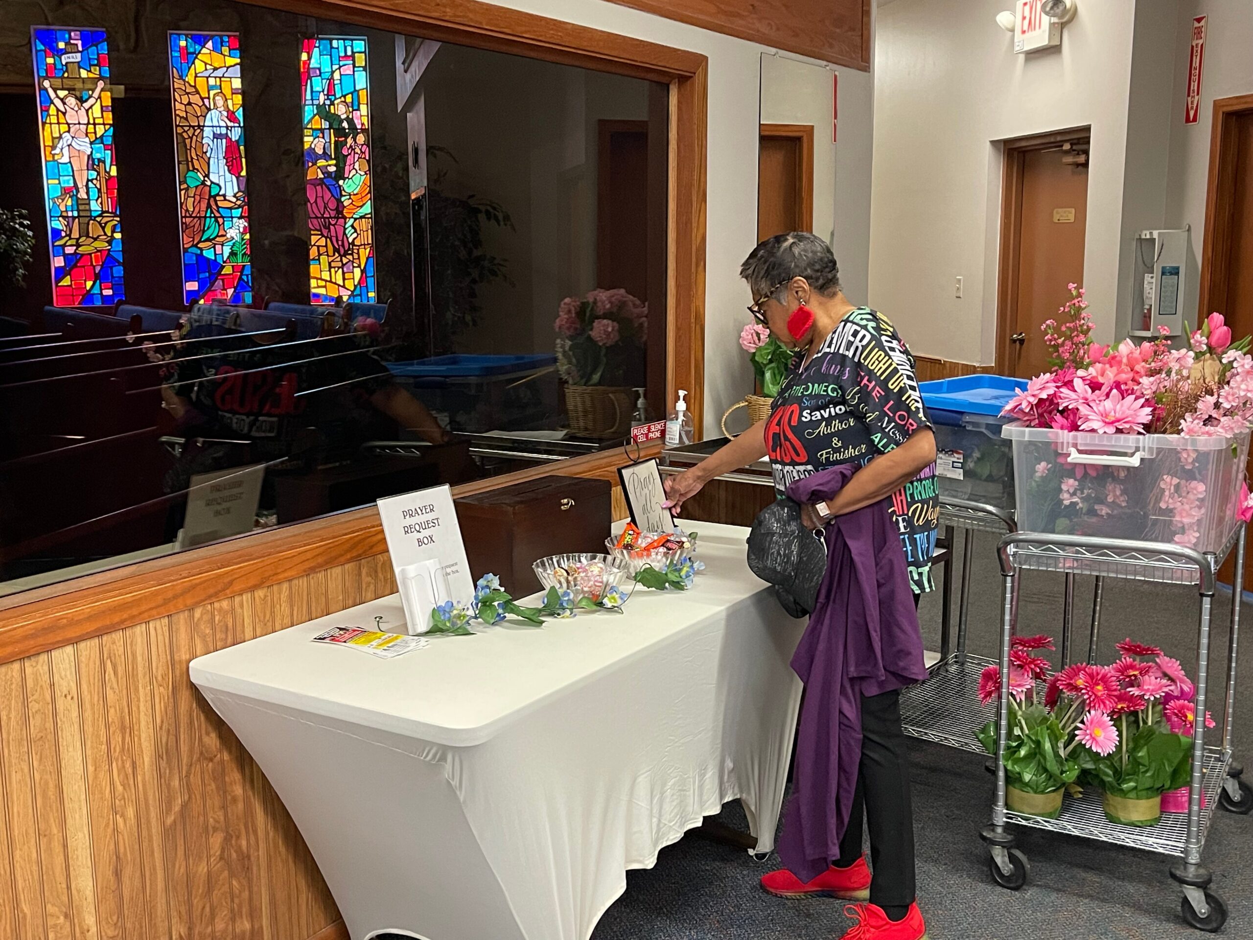 Woman at a church reception desk placing items into a 'Prayer Request Box' with pink flowers nearby and colorful stained glass in the background.