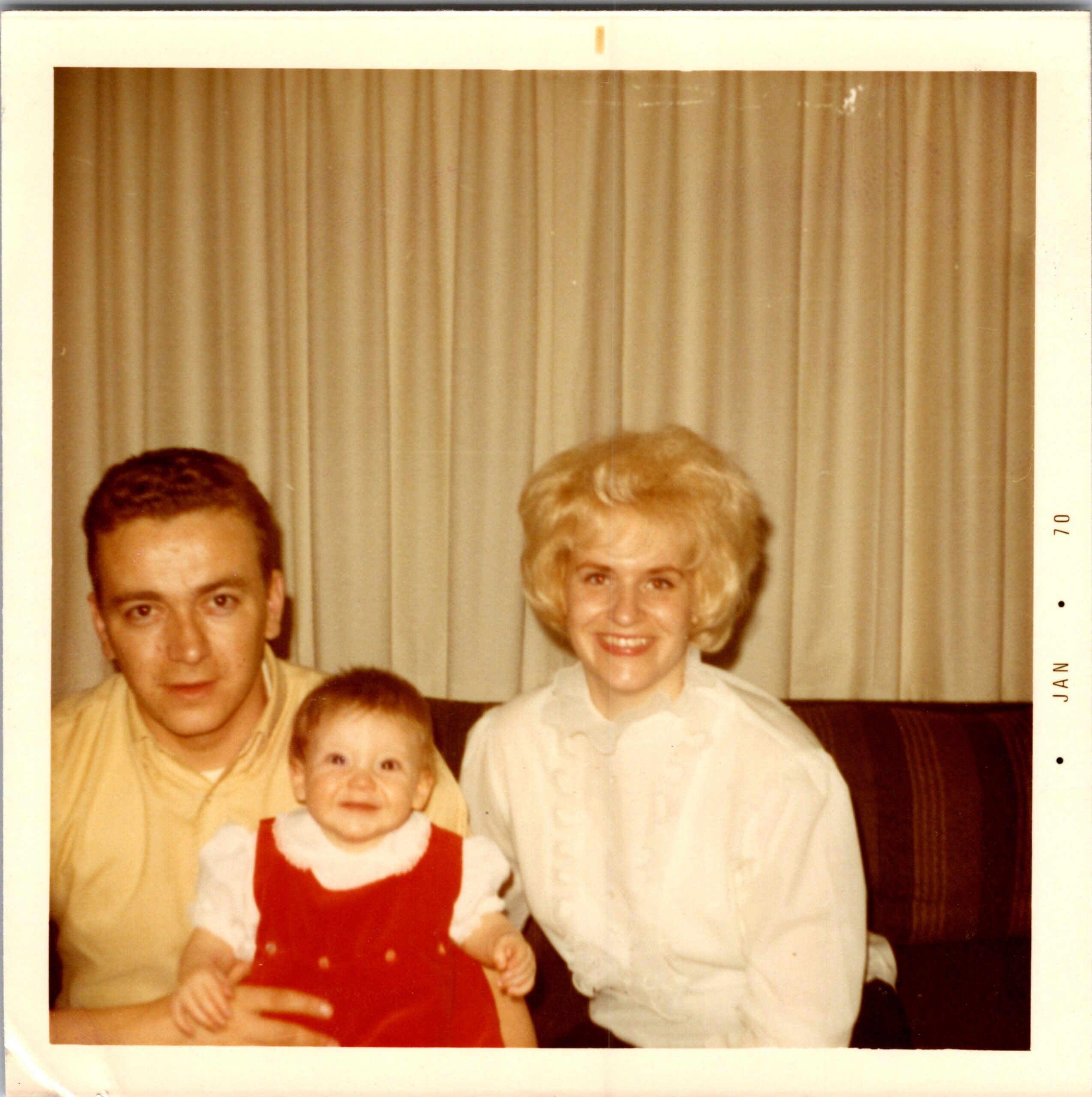 Family portrait indoors with a man on the left, a smiling woman on the right, and a baby in a red dress in the foreground, posed in front of beige curtains.