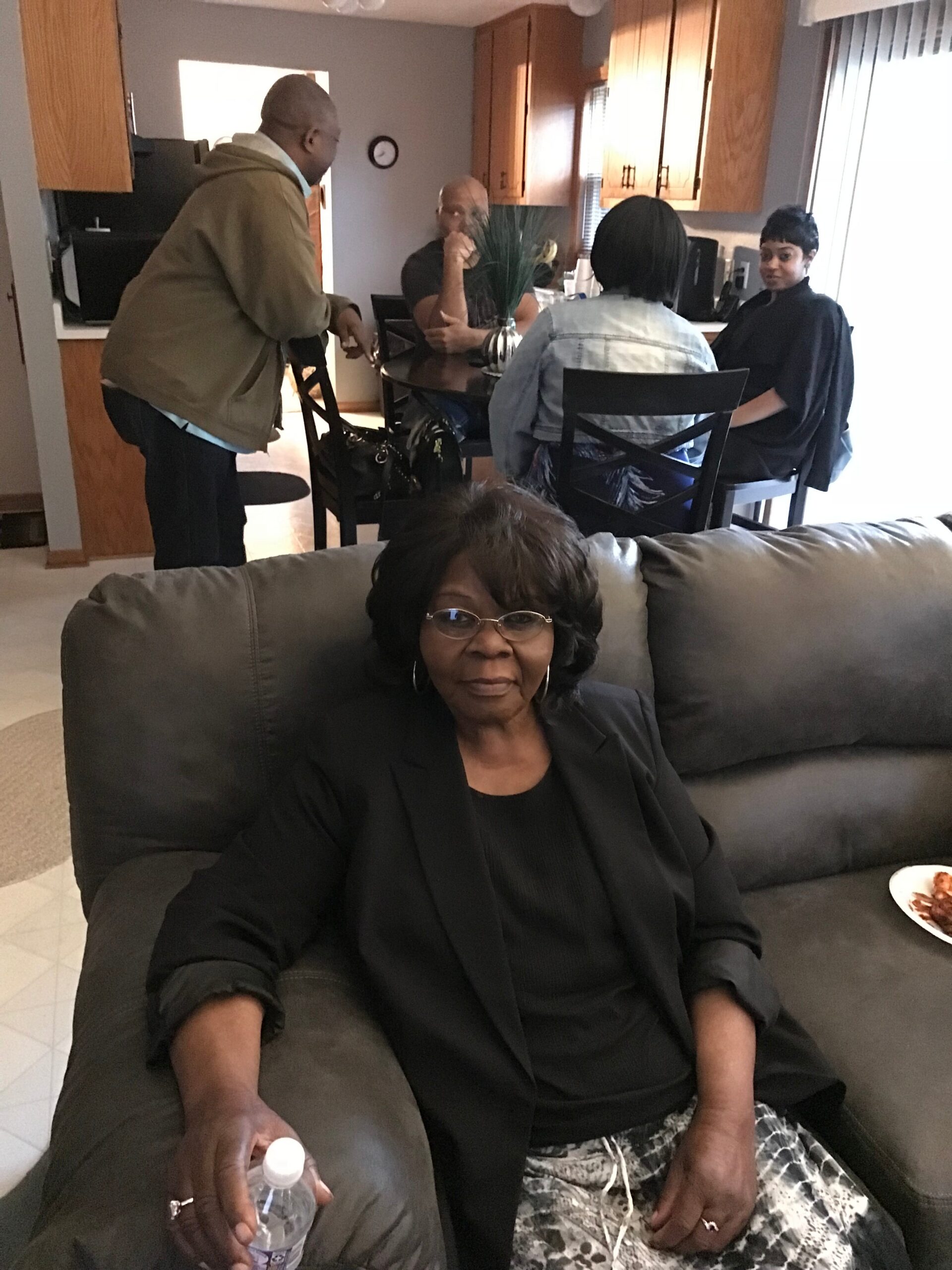 Older woman in foreground sitting on a gray sofa with a water bottle; a small group of four people gathered around a glass dining table in a kitchen.
