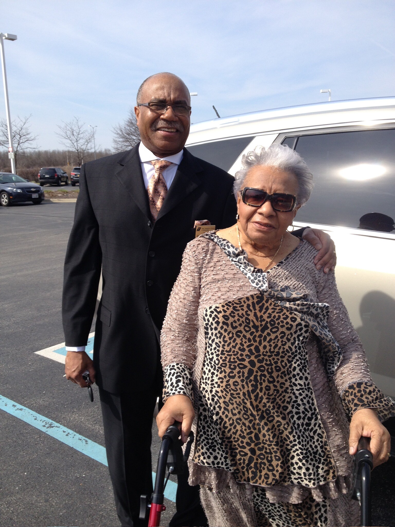 Two smiling elderly people, a man in a suit and a woman in a leopard-print outfit, stand together in a parking lot beside a silver SUV.