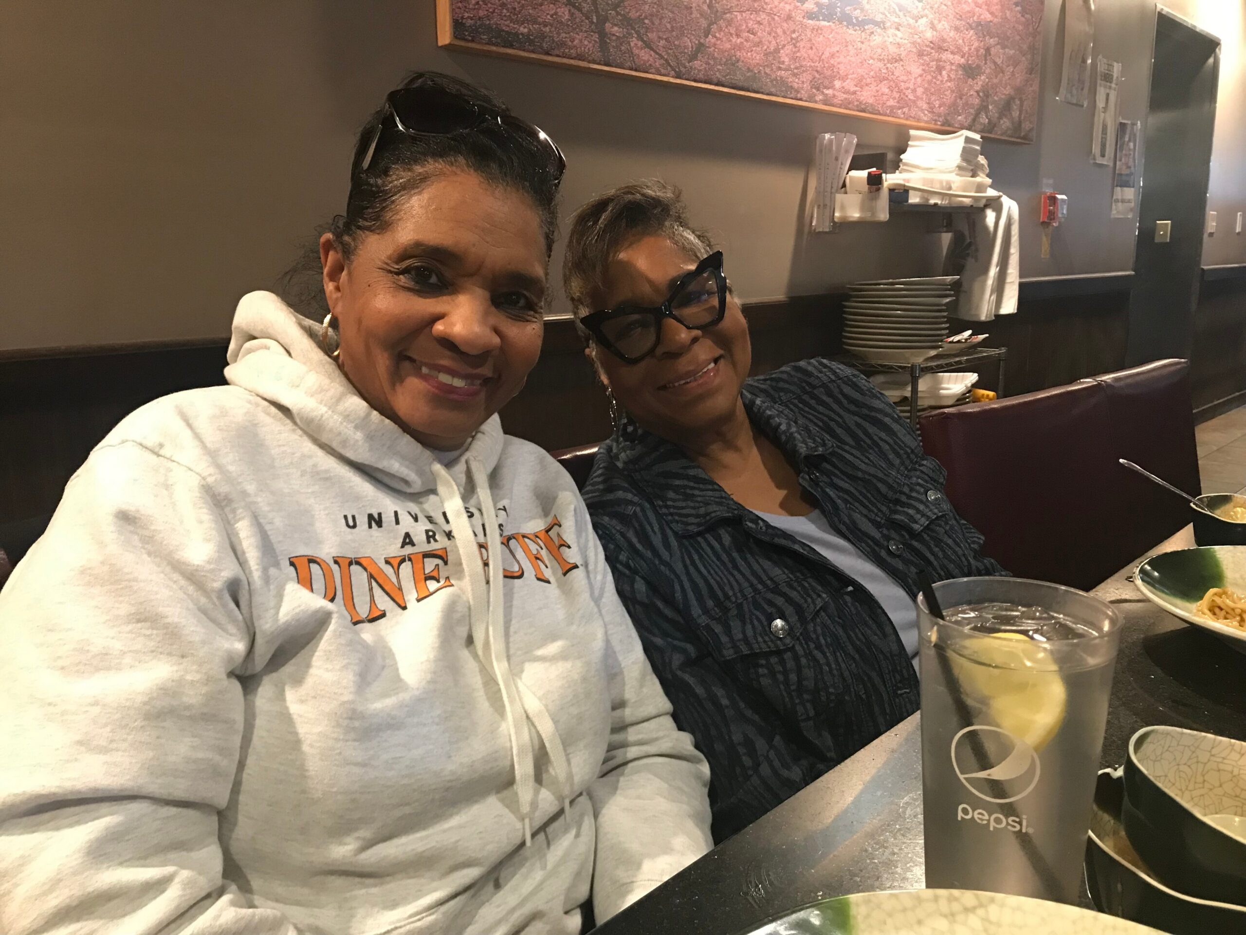 Two women smiling for a selfie in a restaurant, with drinks and plates on the table in front of them.