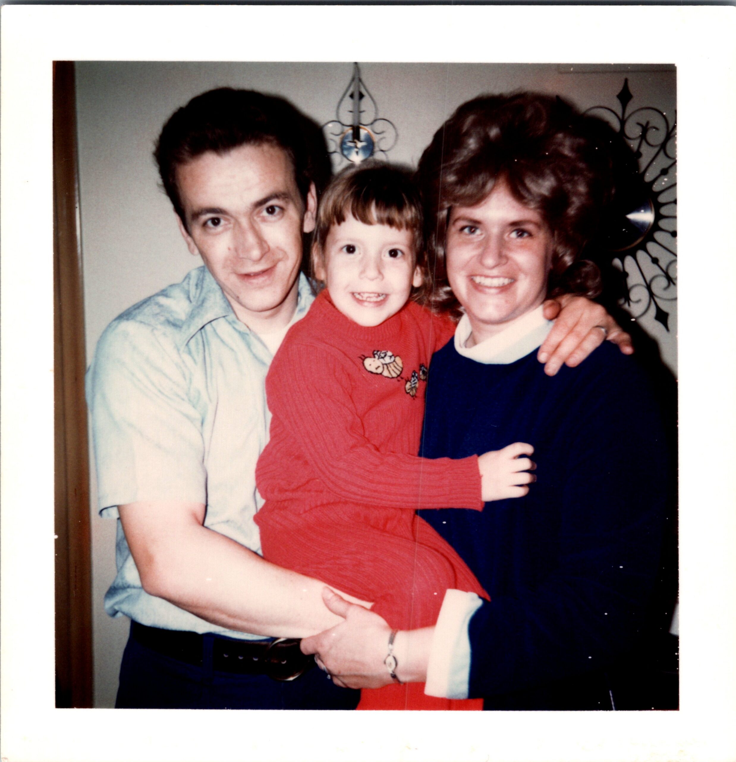 Three people posing together indoors: a man on the left, a smiling child in red in the center, and a woman on the right, with decorative wall art behind them.