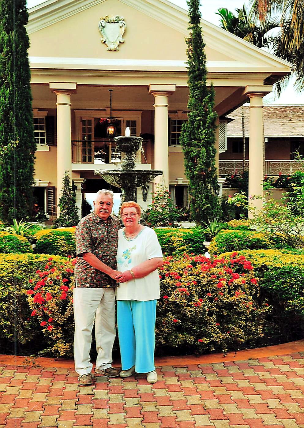 Senior couple standing arm-in-arm in a resort garden with red and yellow flowers and a fountain behind them, in front of a beige columned building.