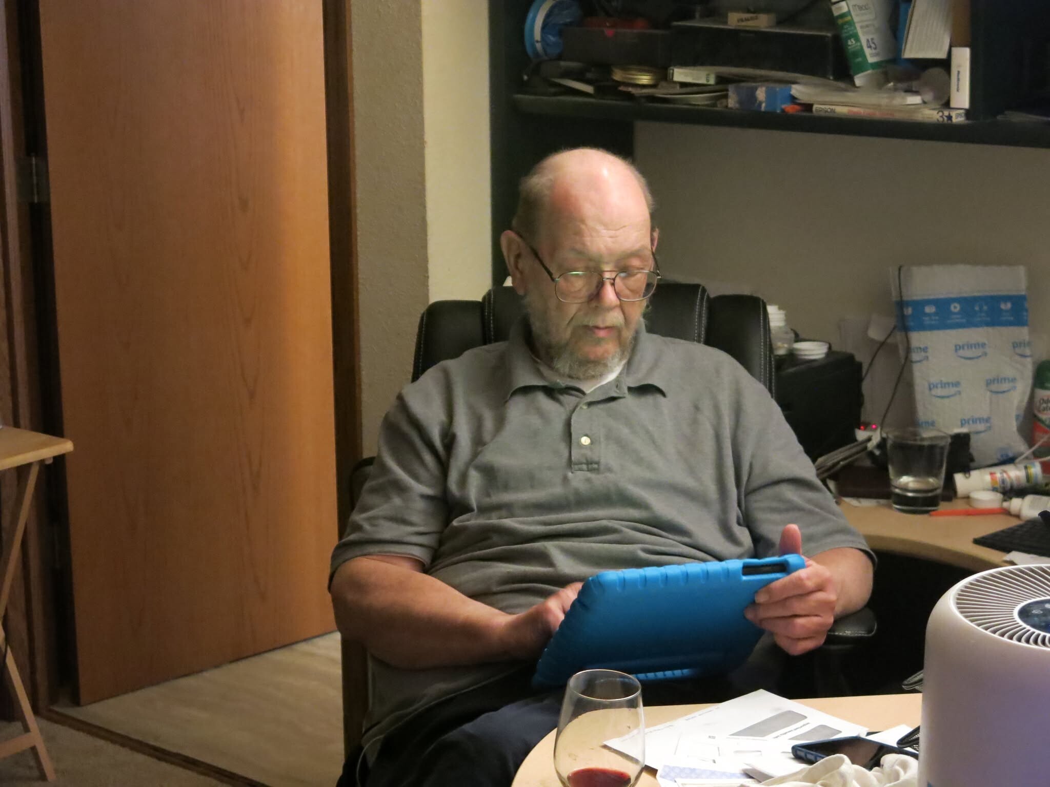 Older man with glasses sits at a desk, looking at a blue tablet or device in his hands in a cluttered office space.