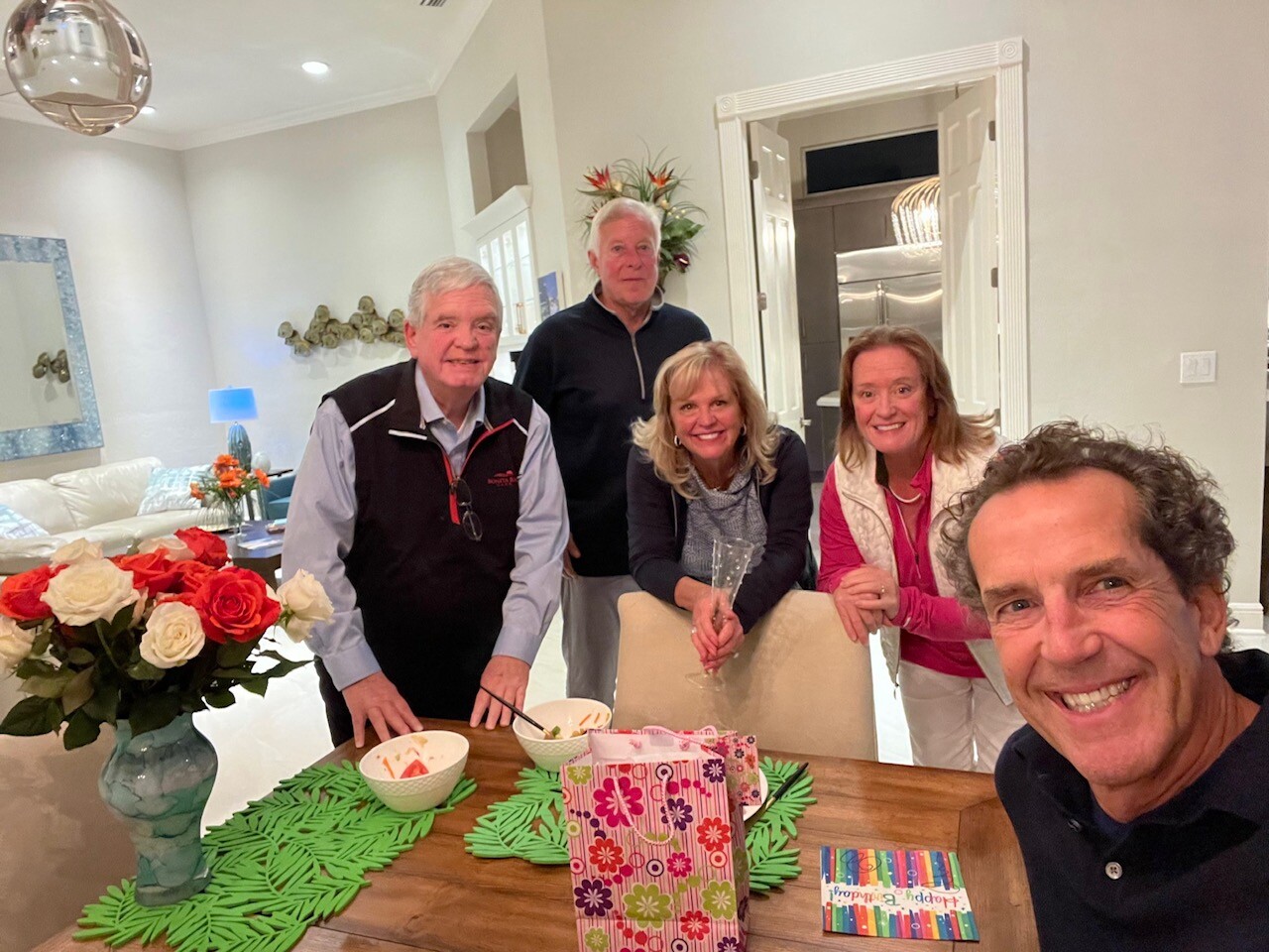 Five adults pose for a selfie around a wooden dining table in a bright living room, with a vase of red and white roses, bowls of snacks, a colorful gift bag, and a striped card on the table.