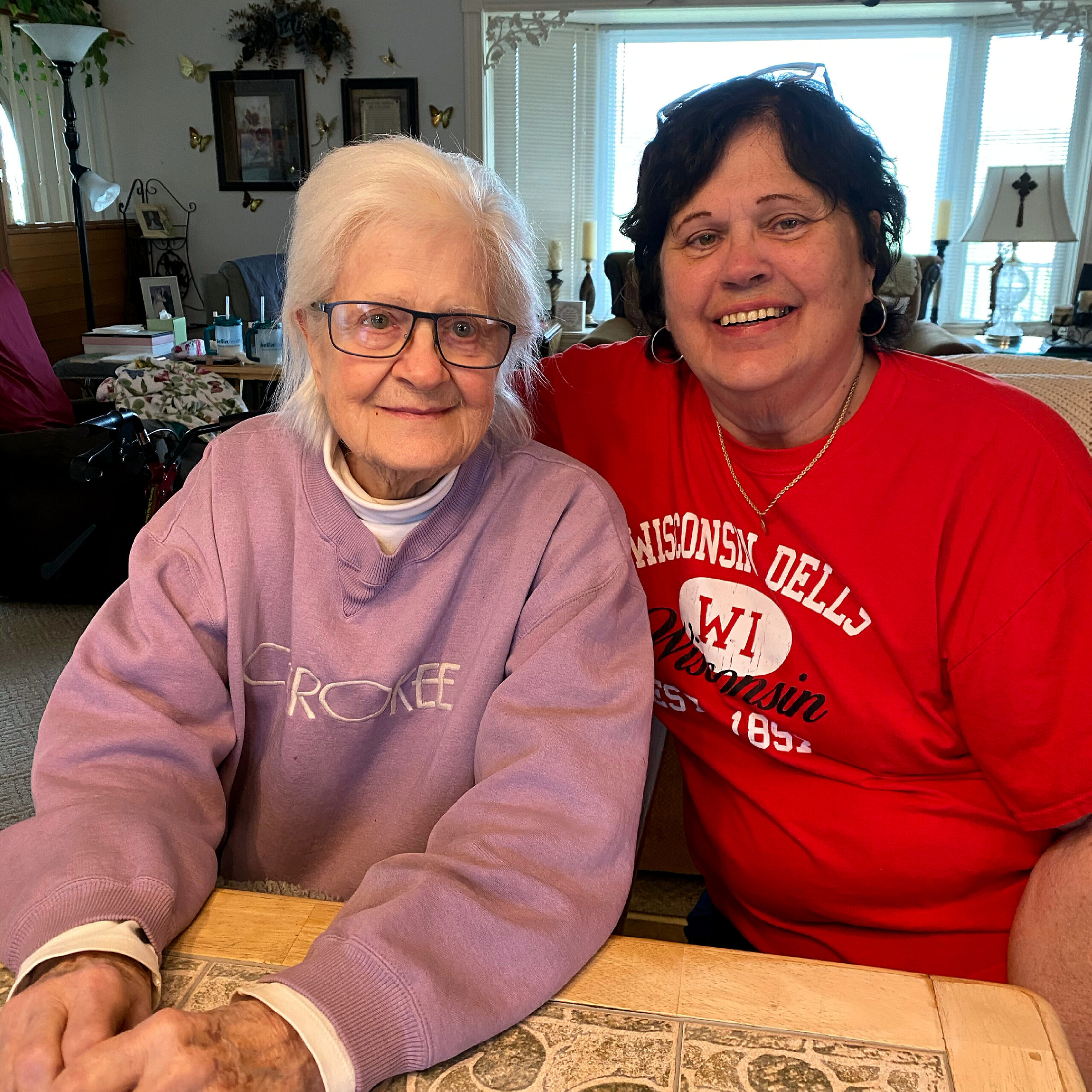 Two smiling women posing for a selfie in a cozy living room; an elderly woman with white hair and glasses sits next to a younger woman in a red shirt.