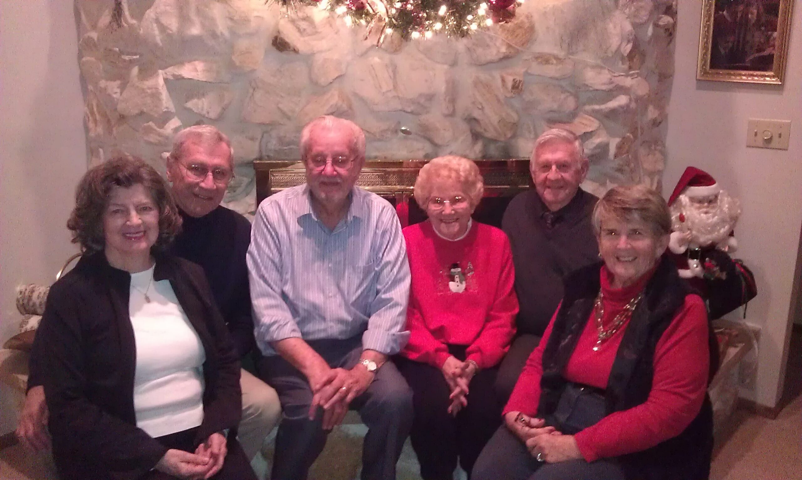 Seven seniors seated together for a holiday photo by a stone fireplace, all smiling at the camera. In the background is festive decor and a Santa prop on the right.