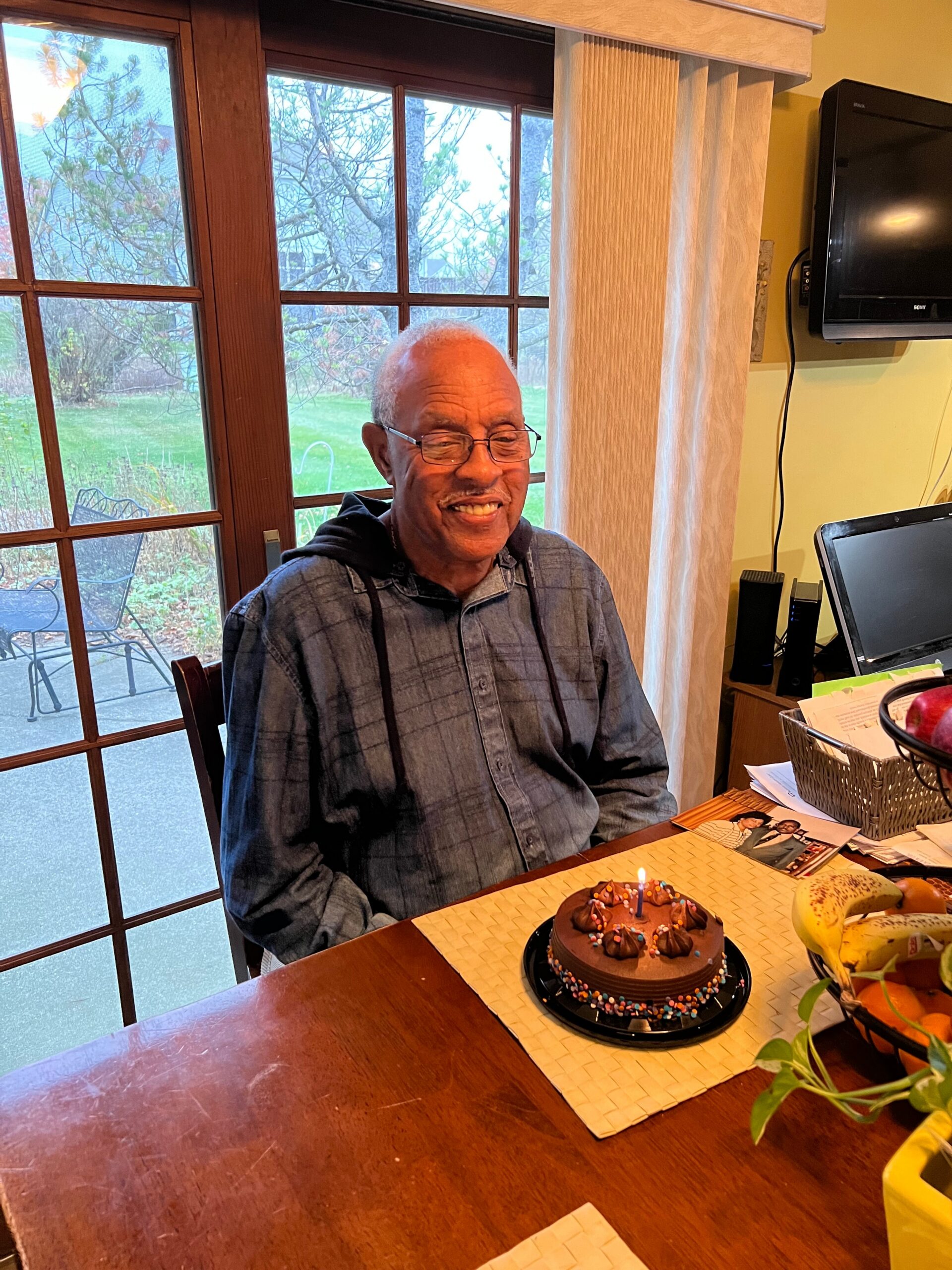 An elderly man with glasses sits at a dining table, smiling, in front of a chocolate birthday cake with candles and sprinkles.