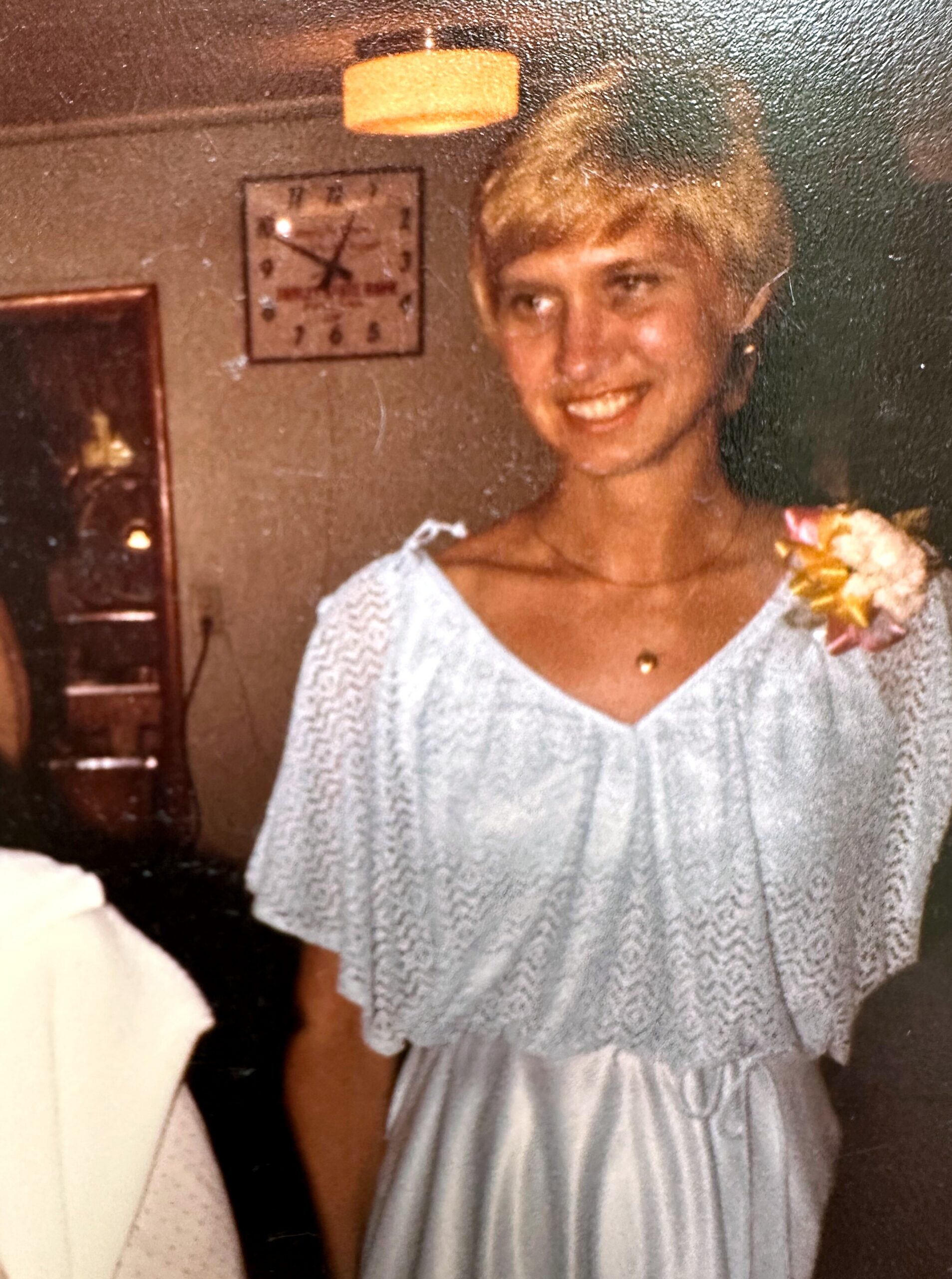 Smiling woman in a light lace dress with a flower corsage at a party, indoors with a wall clock behind her