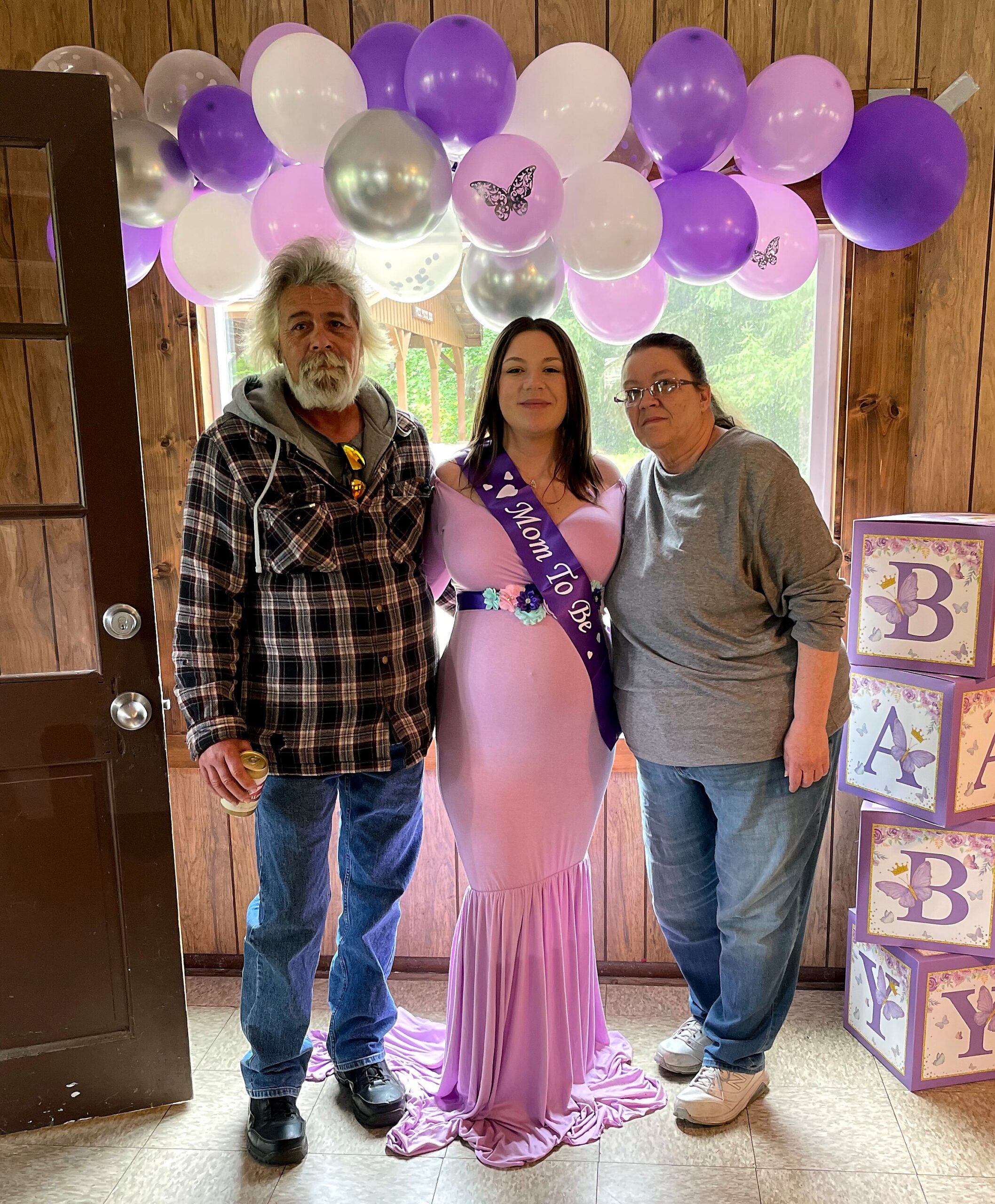 Pregnant woman in a pink gown with a 'Mom to Be' sash stands between two adults under a purple-pink balloon arch in a wood-paneled room.