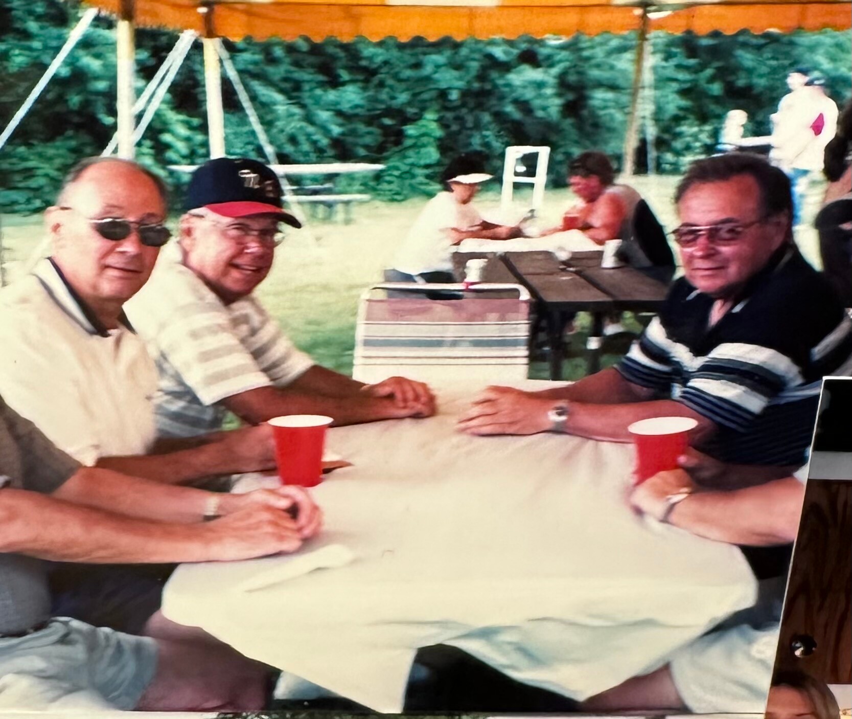 Three men sit at a picnic table outdoors under a pavilion, chatting with red plastic cups in front of them.