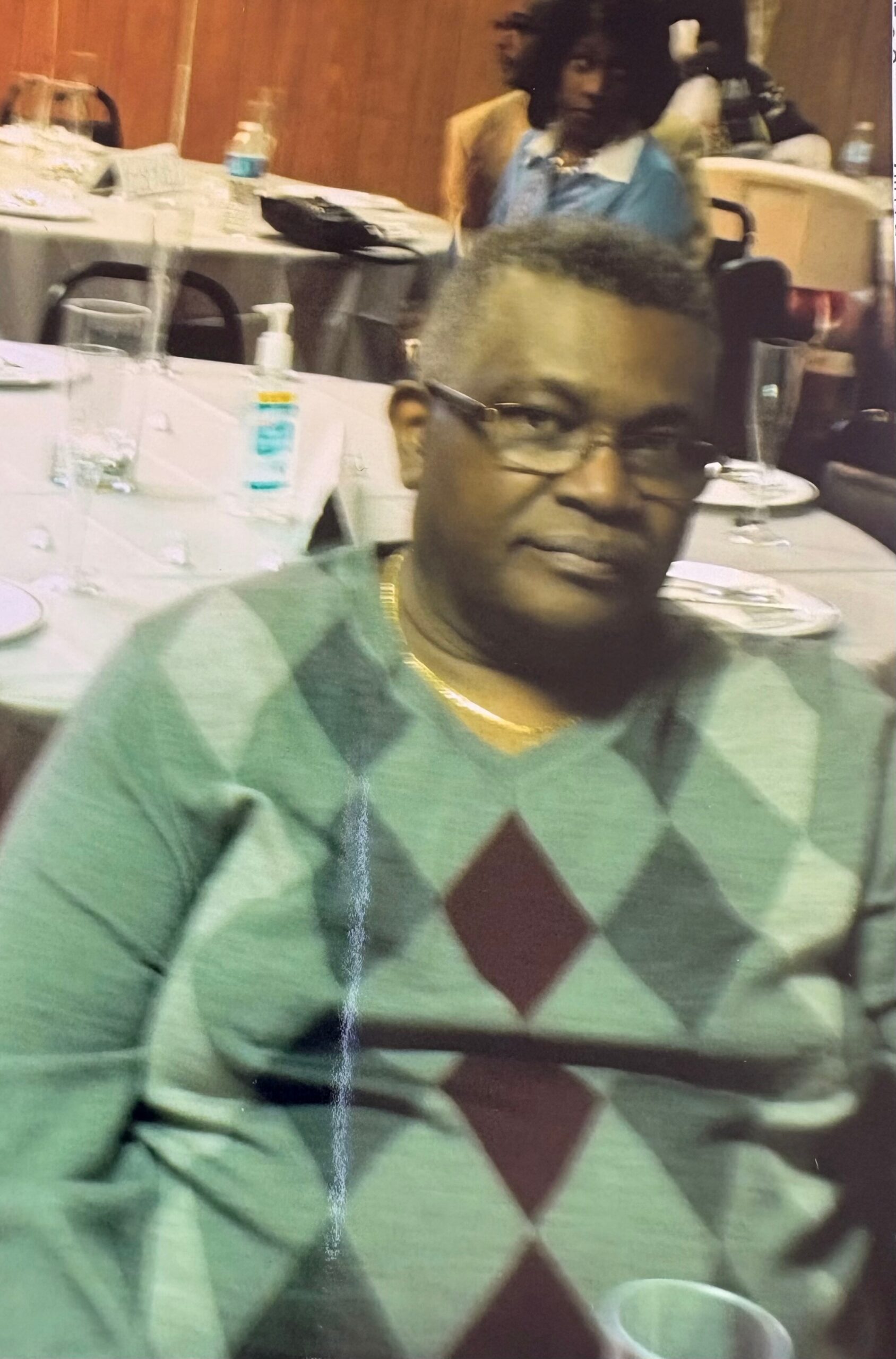 Close-up of a person with short dark hair and glasses wearing a green argyle sweater, seated at a dining table with plates and bottles in the background.