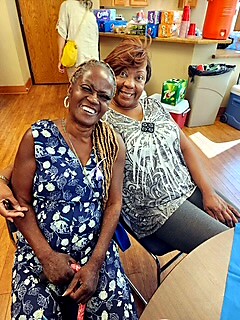 Two smiling women sit side by side in a bright community room, posing for a photo with a snack table in the background.