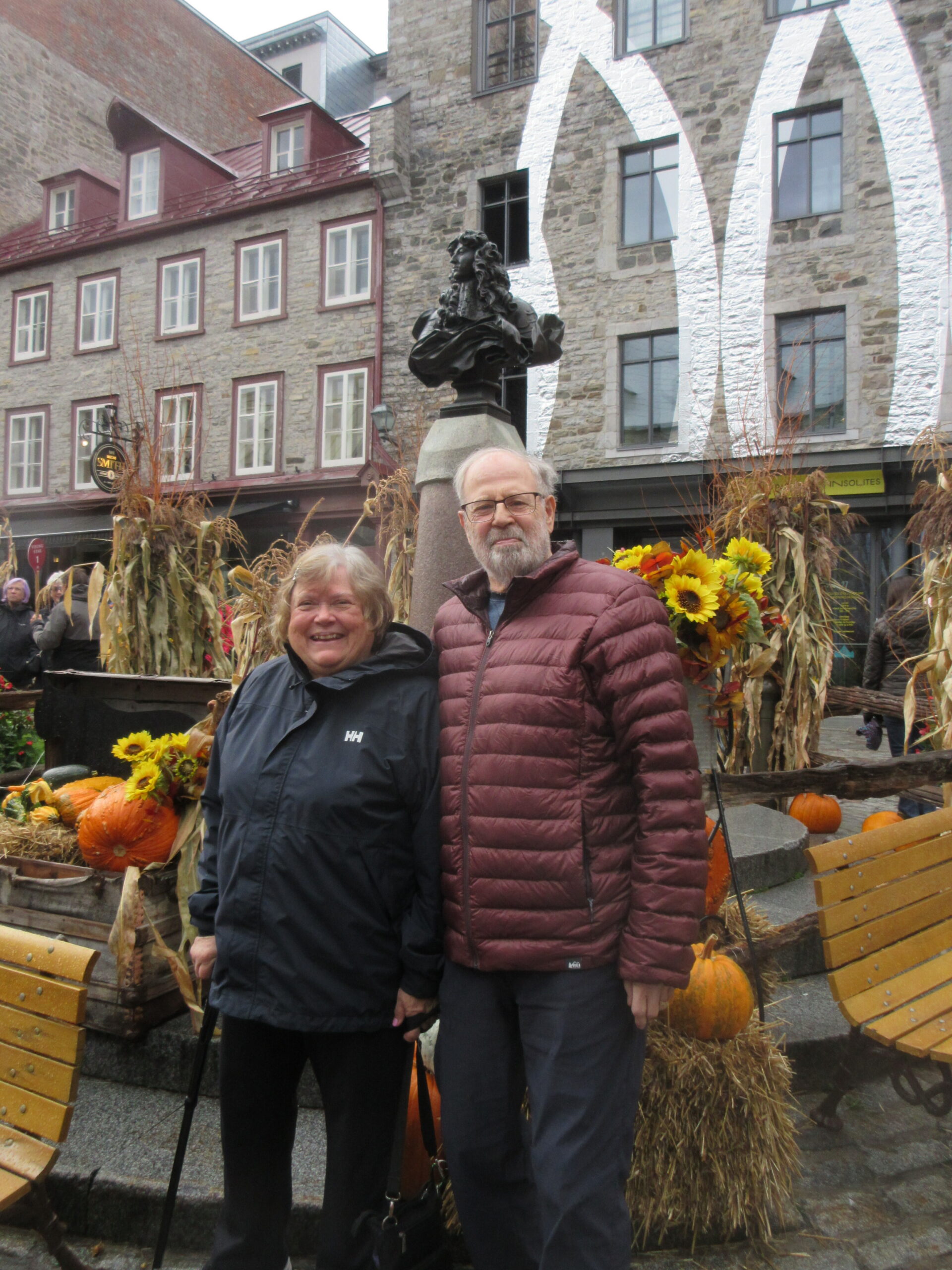 Smiling elderly couple in jackets posing in front of a statue on a stone pedestal amid an autumn harvest display with pumpkins nearby.