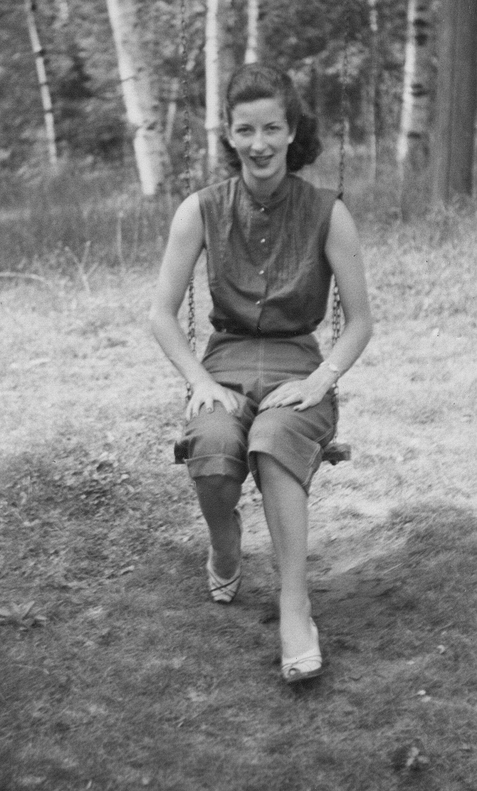 Woman sits on a swing in a forest, smiling at the camera.