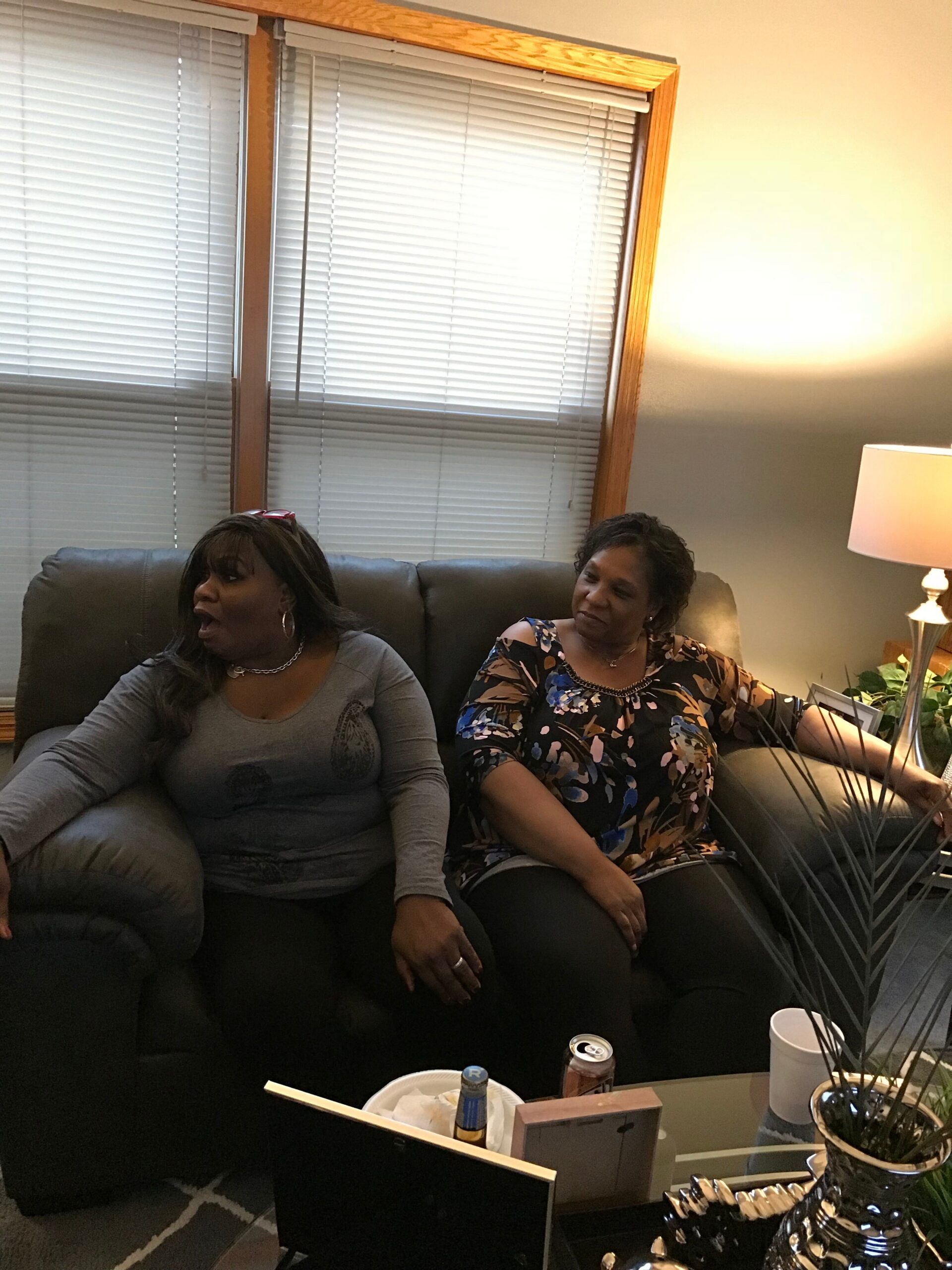 Two women sit on a dark sofa in a living room, chatting by a window with blinds and a lit table lamp nearby.