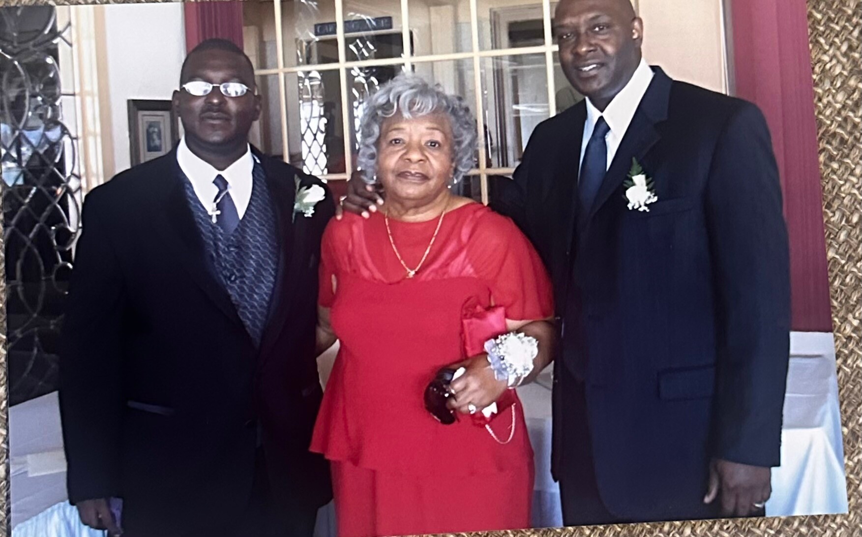 Three adults in formal attire pose for a photo: two men in suits flank an elderly woman in a red dress with a corsage and clutch, at a celebration.