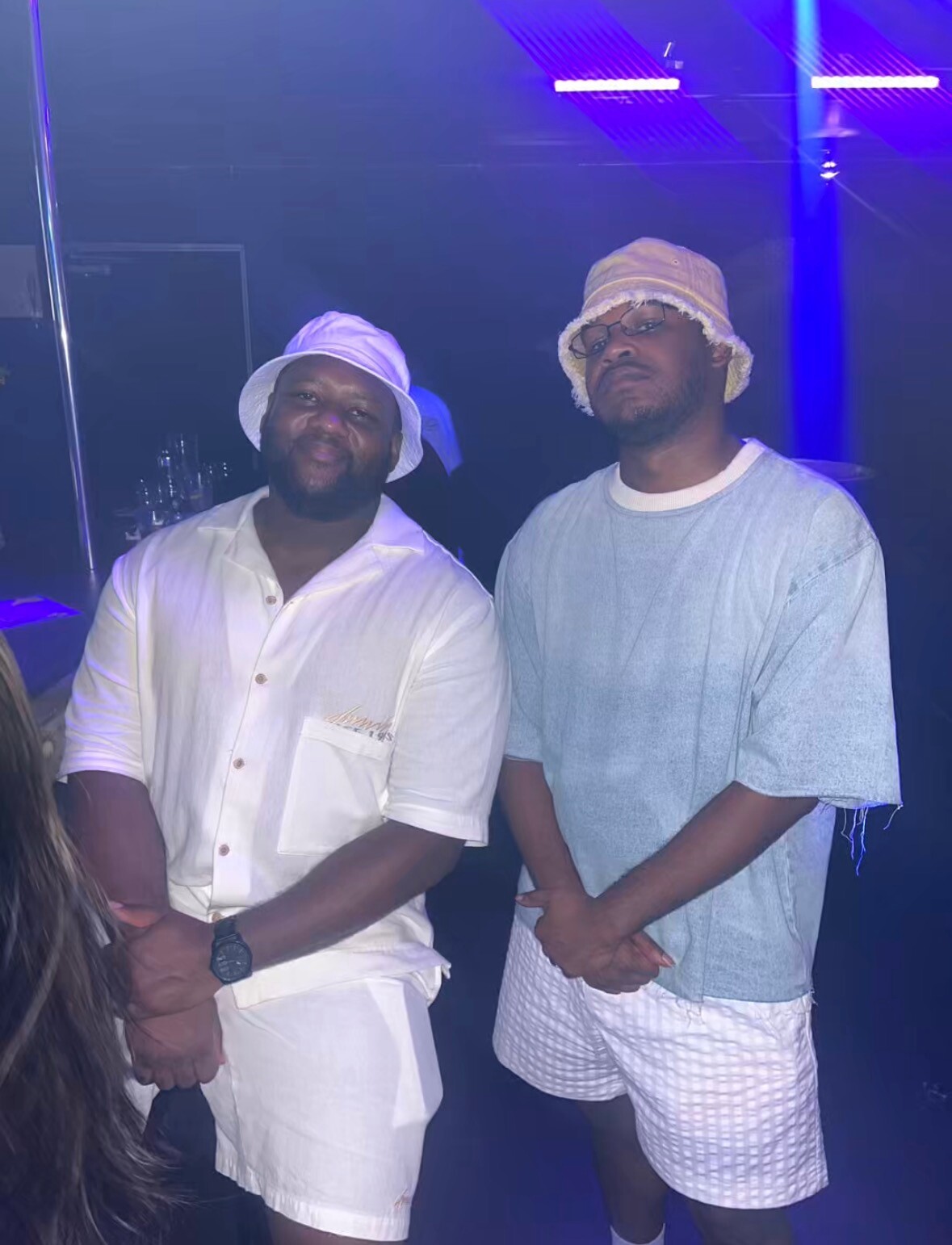 Two men posing at a party under blue/purple lighting; one in a white shirt and hat, the other in a light denim shirt and bucket hat.