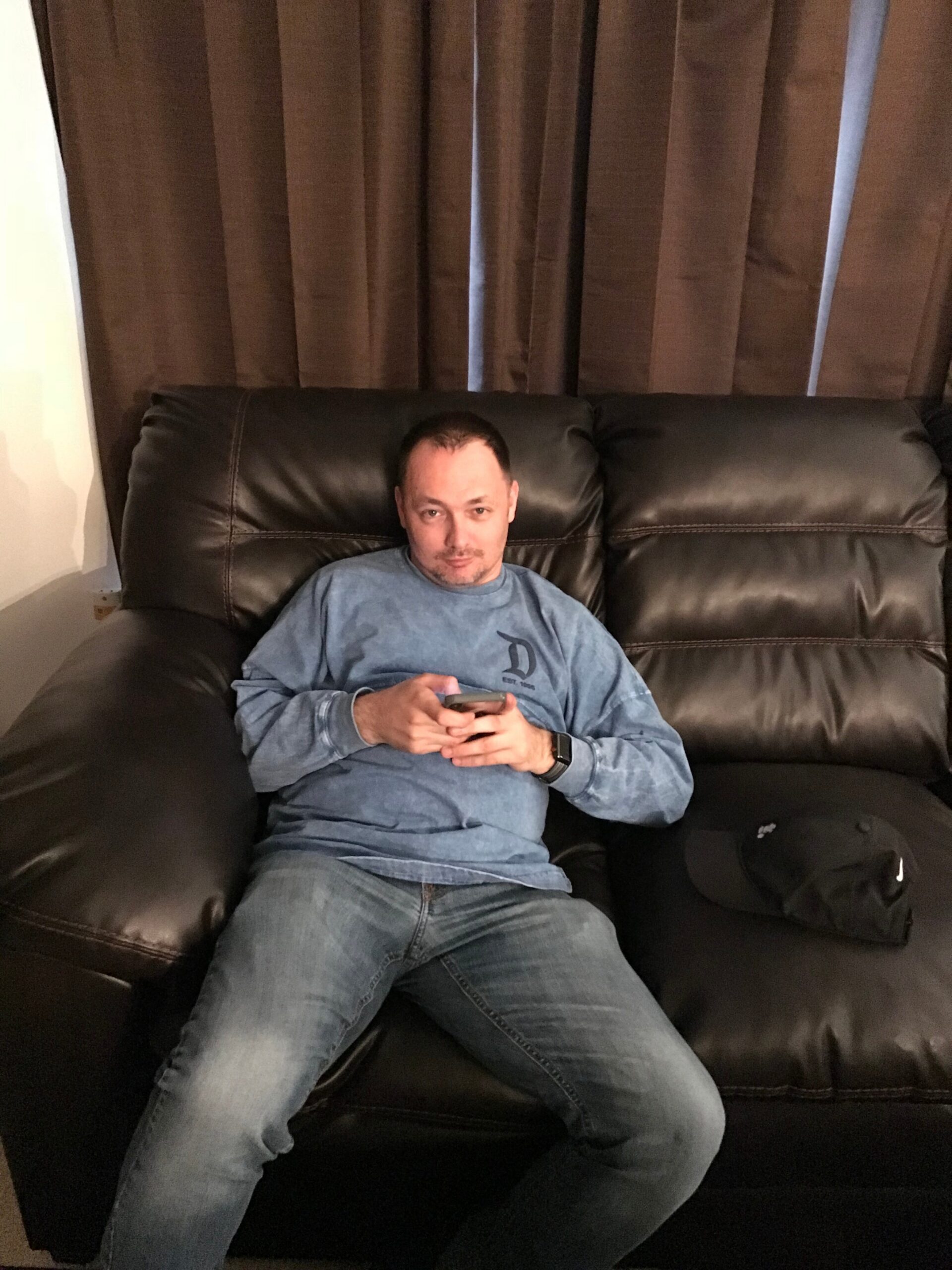 Man lounging on a dark leather sofa, wearing a blue long-sleeve shirt and jeans, looking at a smartphone in his hands with brown curtains in the background.