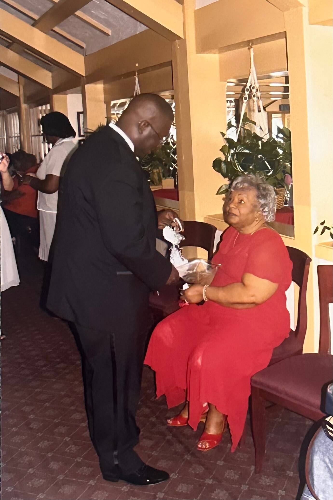 A man in a suit hands a small package to an elderly woman in a red dress at an indoor event, others in background.