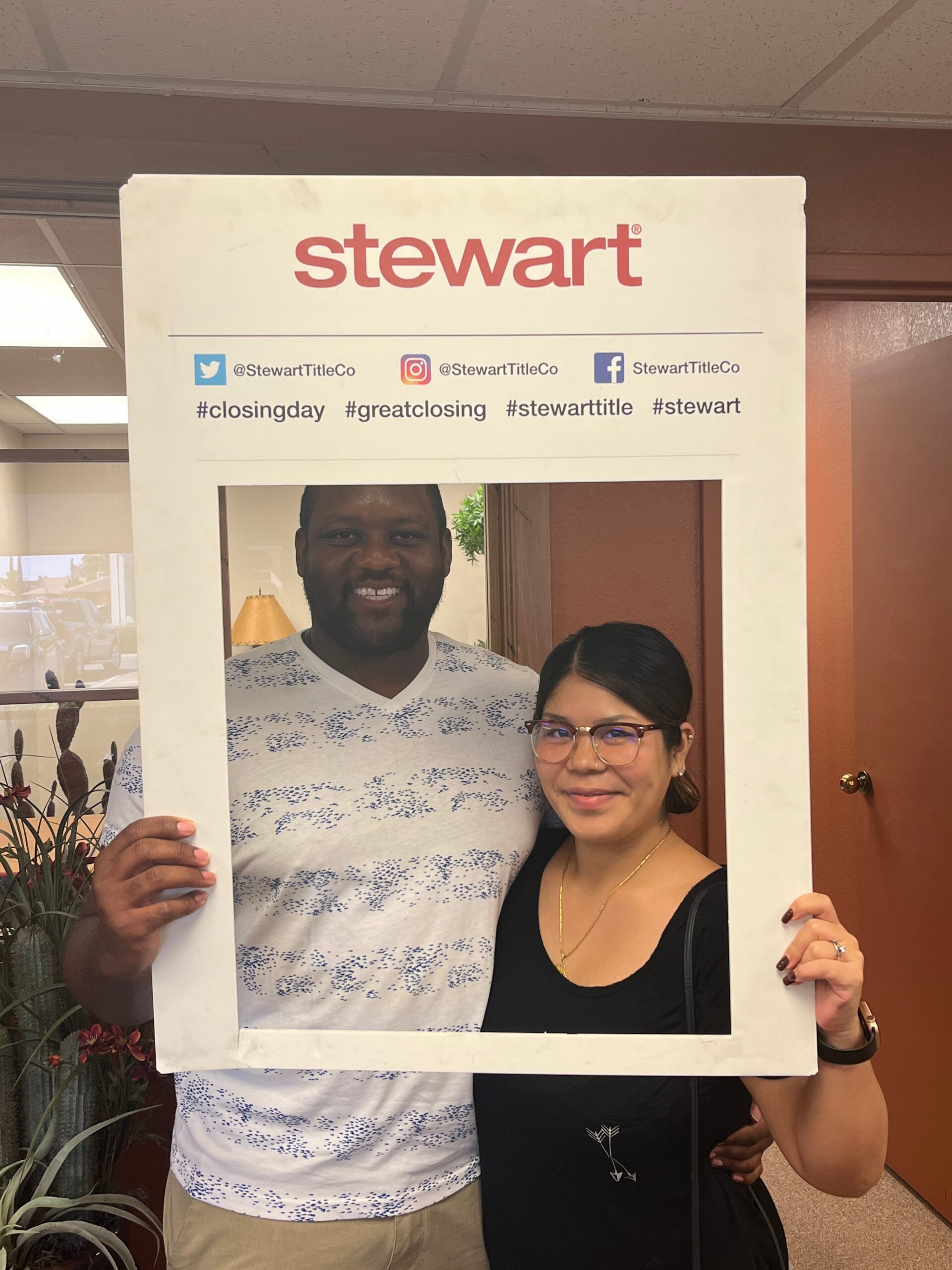 Two people pose together inside a white cardboard photo frame with 'stewart' branding and social icons behind them in a store.