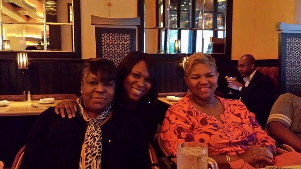 Three women sit together at a restaurant booth, smiling for the camera; warm lighting and decorative panels behind them.