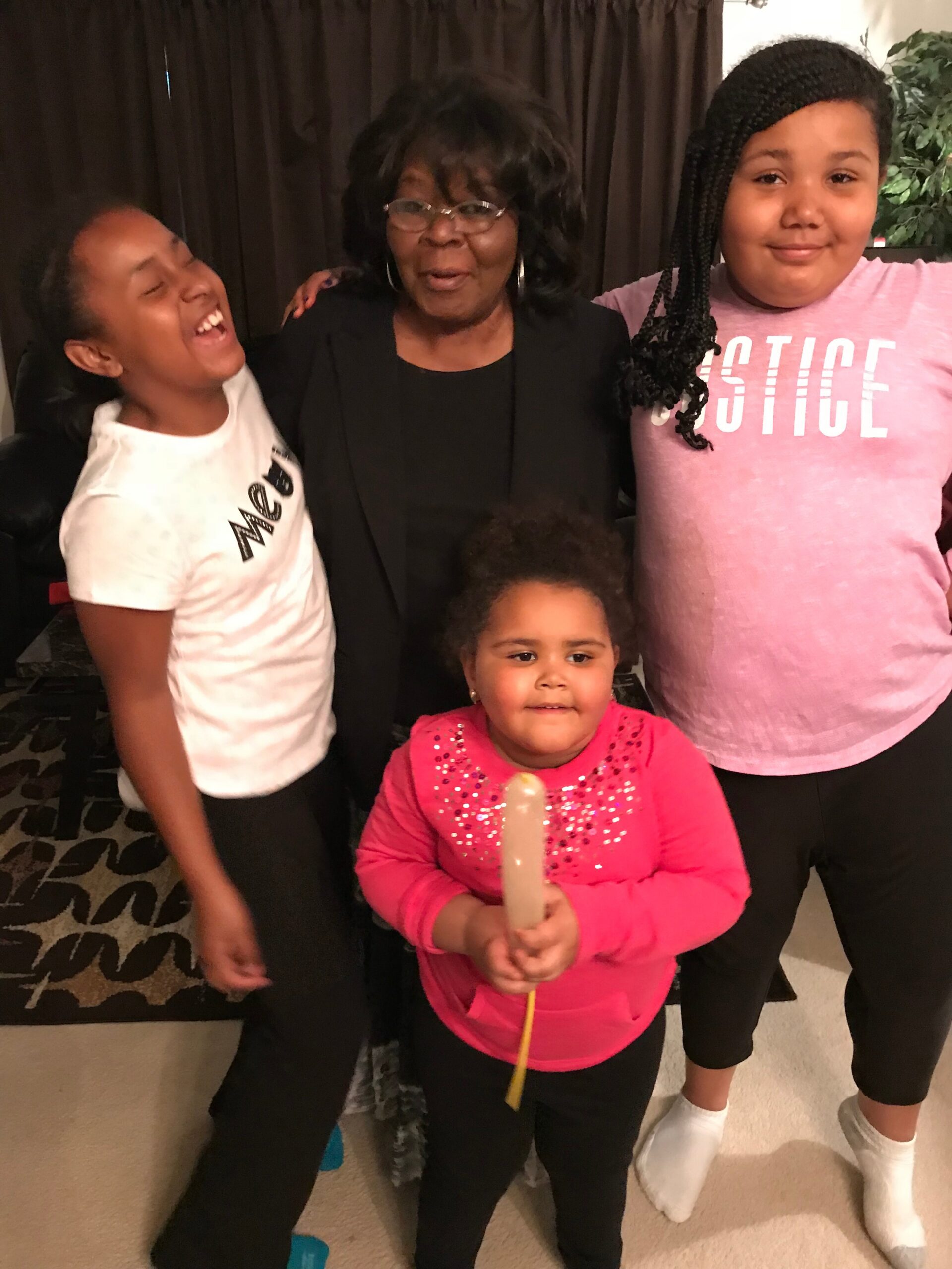 Smiling woman in a black blazer surrounded by three children posing indoors.