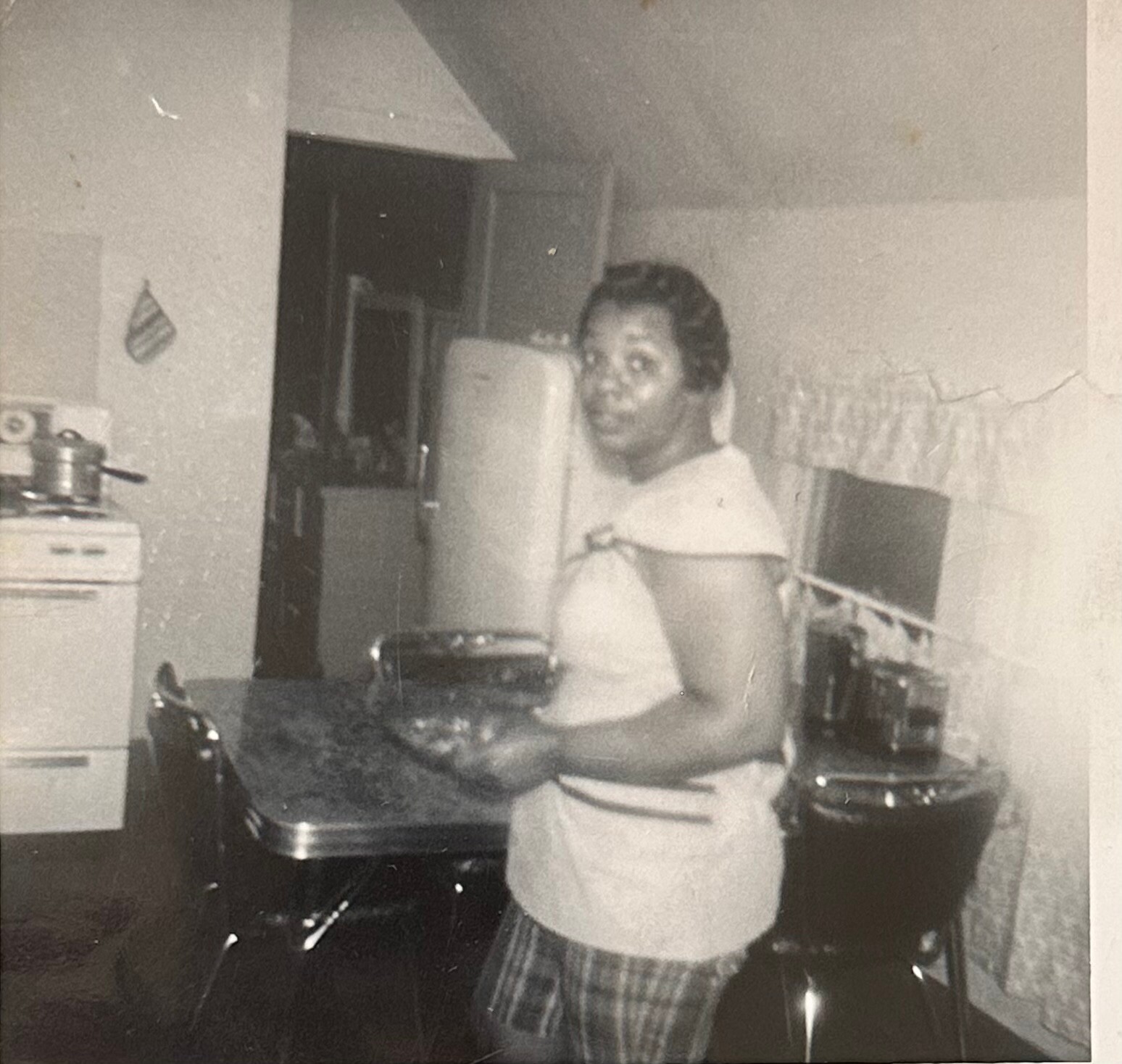 African American woman standing in a mid-century kitchen, wearing a light blouse and plaid pants, looking at the camera with appliances in the background.
