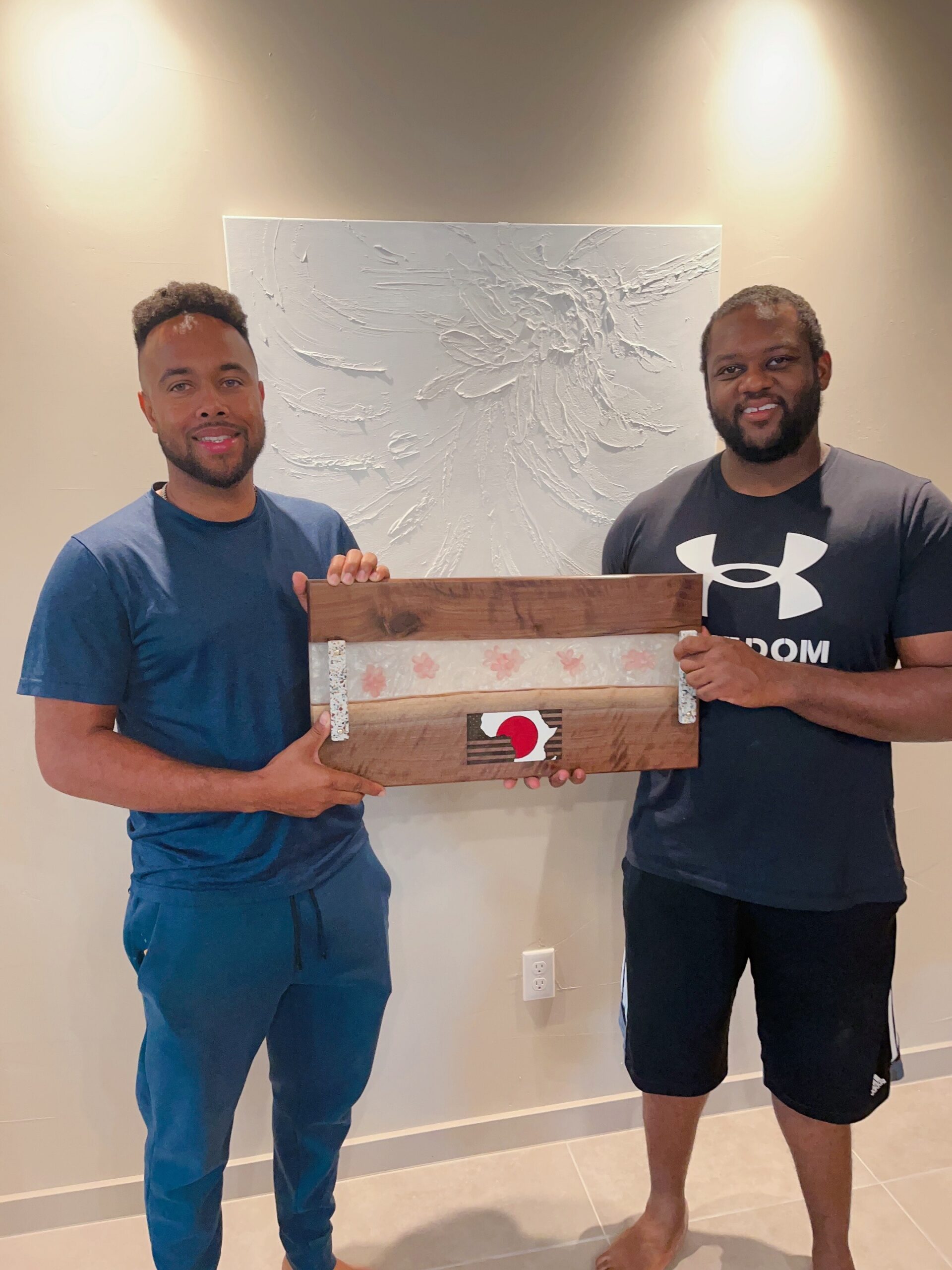 Two men indoors, smiling, holding a wooden sign in front of a textured white wall art piece behind them.