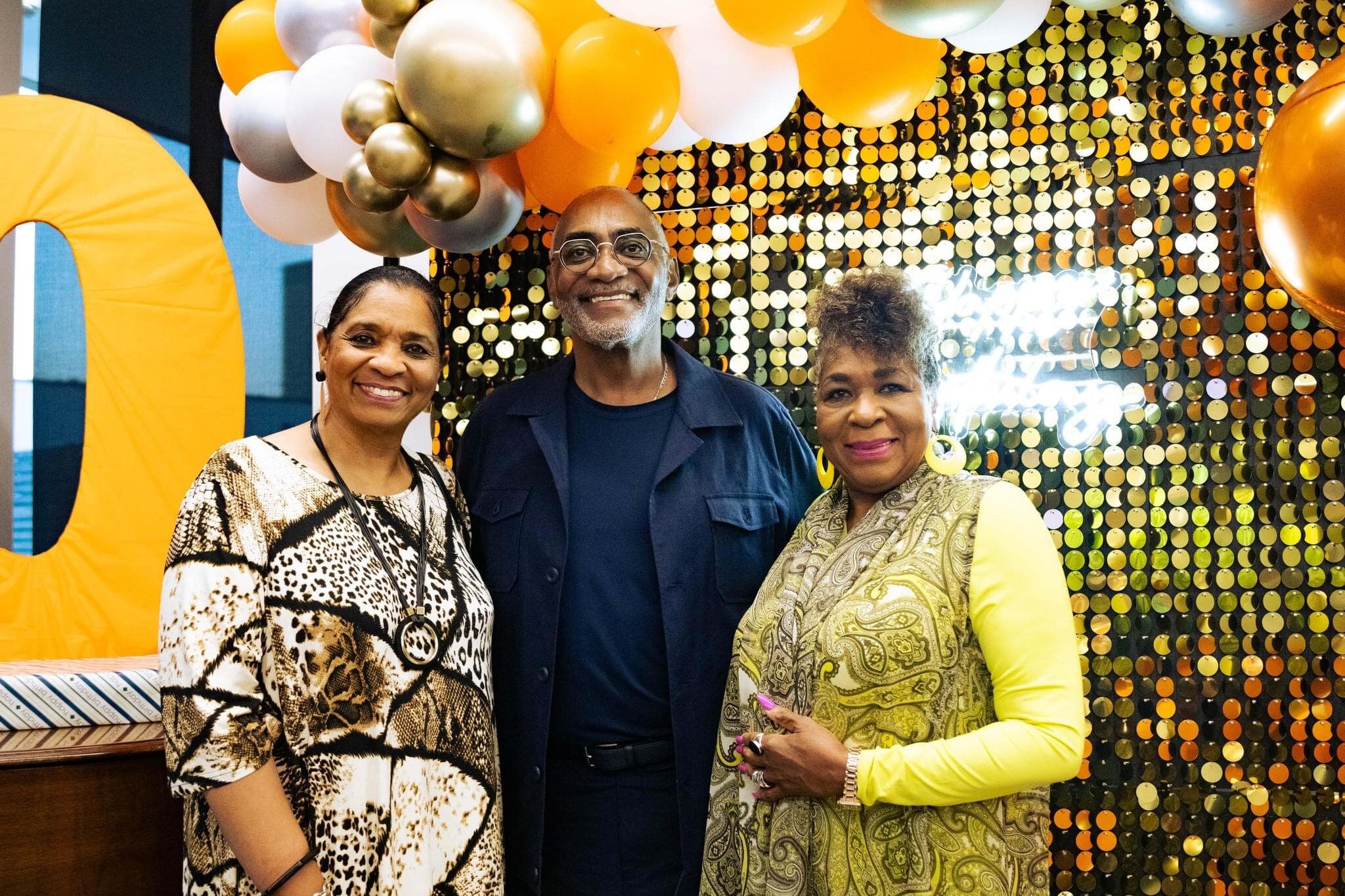 Three smiling adults posing at a celebration with gold balloons and a gold sequin backdrop.