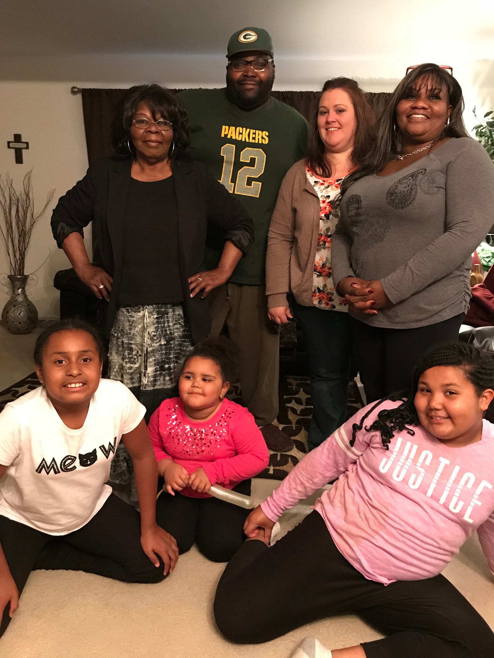 Four adults stand behind several smiling children seated on a carpet in a living room; a man wears a green Packers jersey with number 12, flanked by two women in casual attire and a woman in a black blazer. A wall cross and decorative vase are visible in the background.