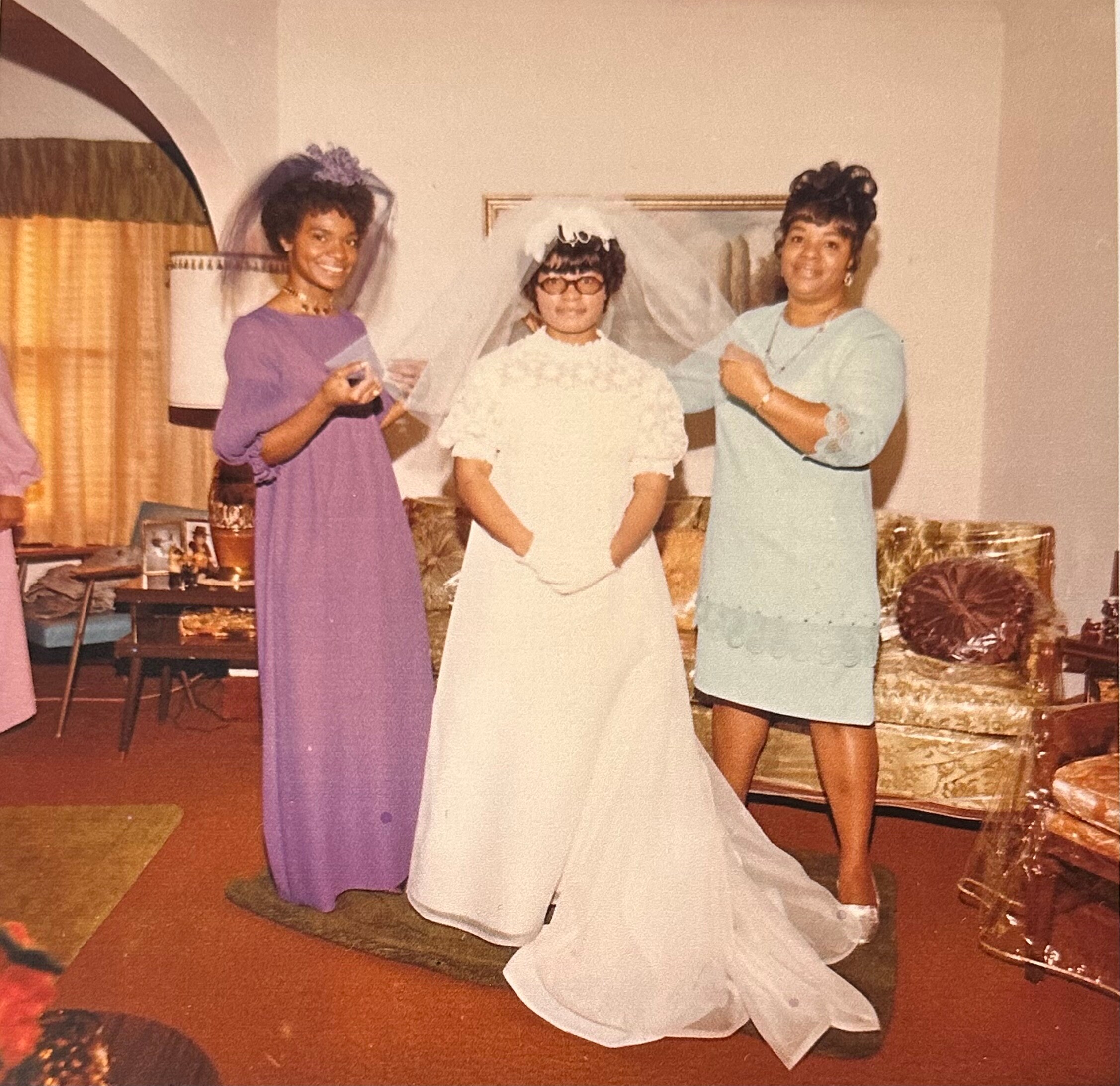 Three women posing at a wedding: the bride in a white gown and veil stands with two bridesmaids in purple and light blue dresses indoors.