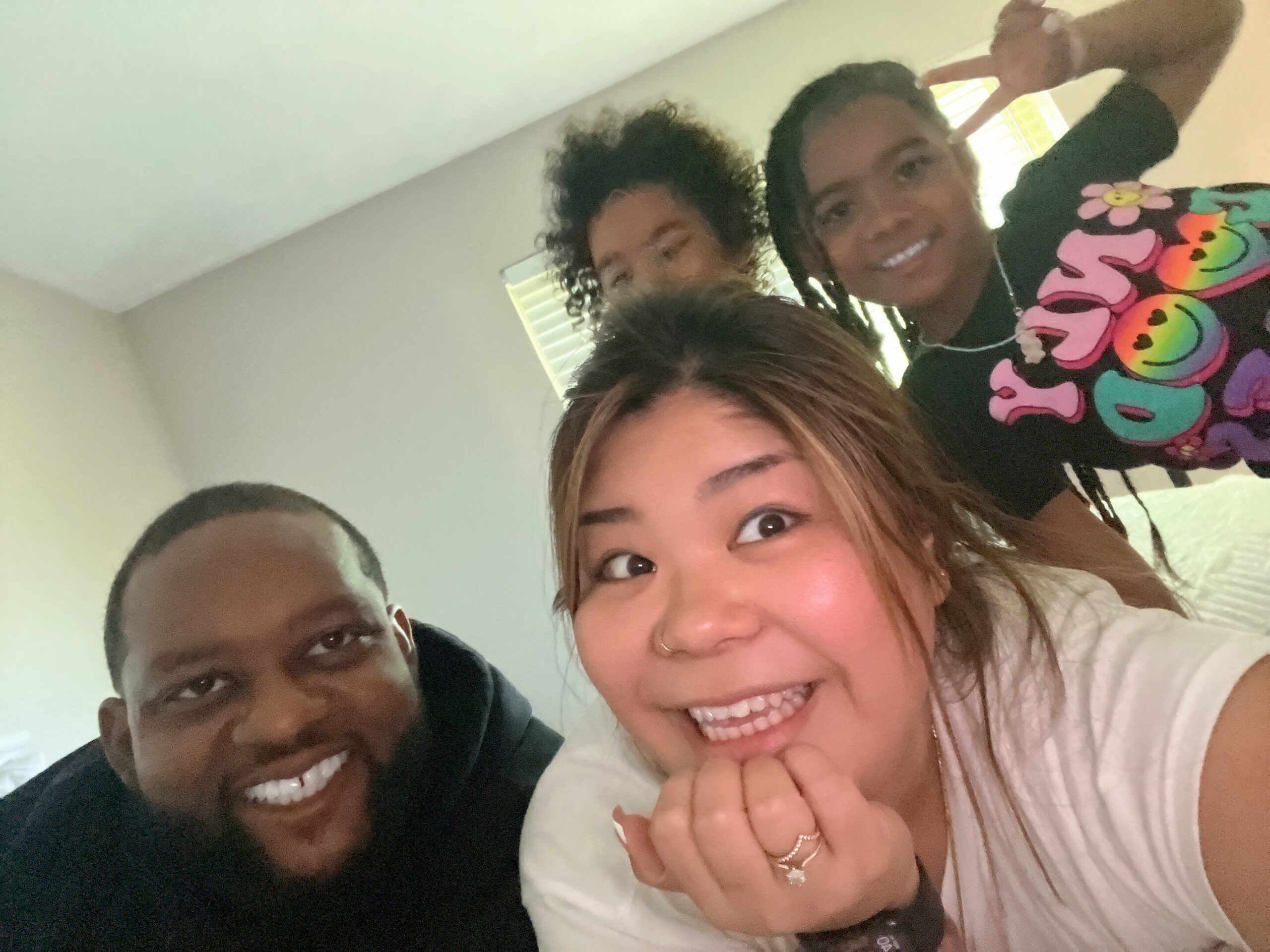 Smiling group selfie indoors with four friends; foreground woman with hands near chin, others in background making playful expressions and peace sign gesture.