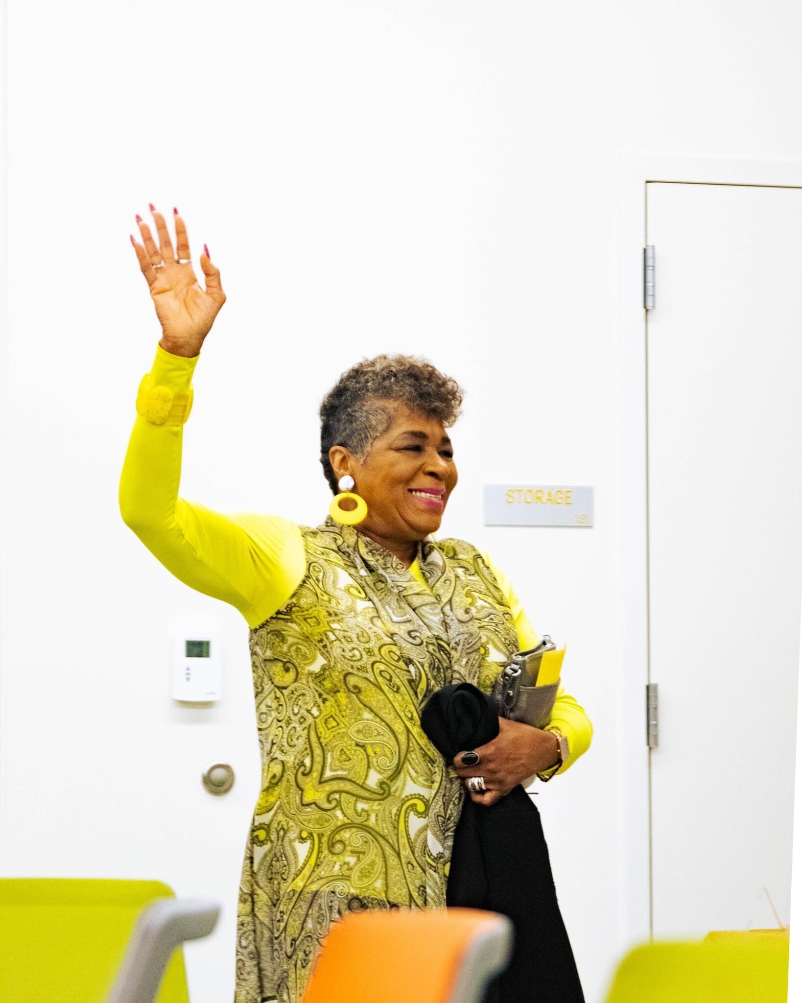 Smiling woman in a bright yellow outfit waves hello indoors, standing near a door with a storage sign in the background.
