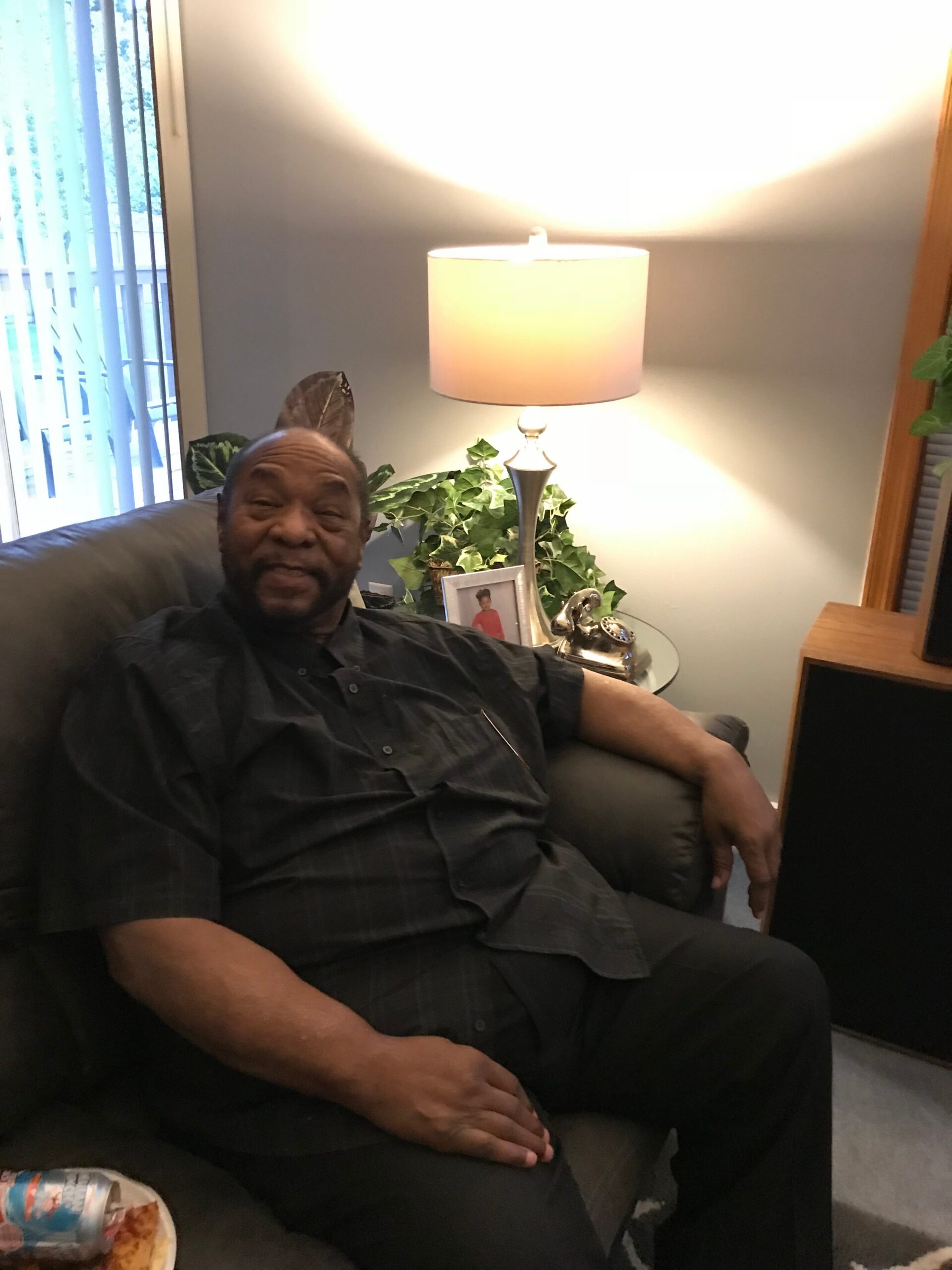 Older man relaxing on a dark sofa in a living room, with a lit table lamp, plants, and a framed photo nearby.