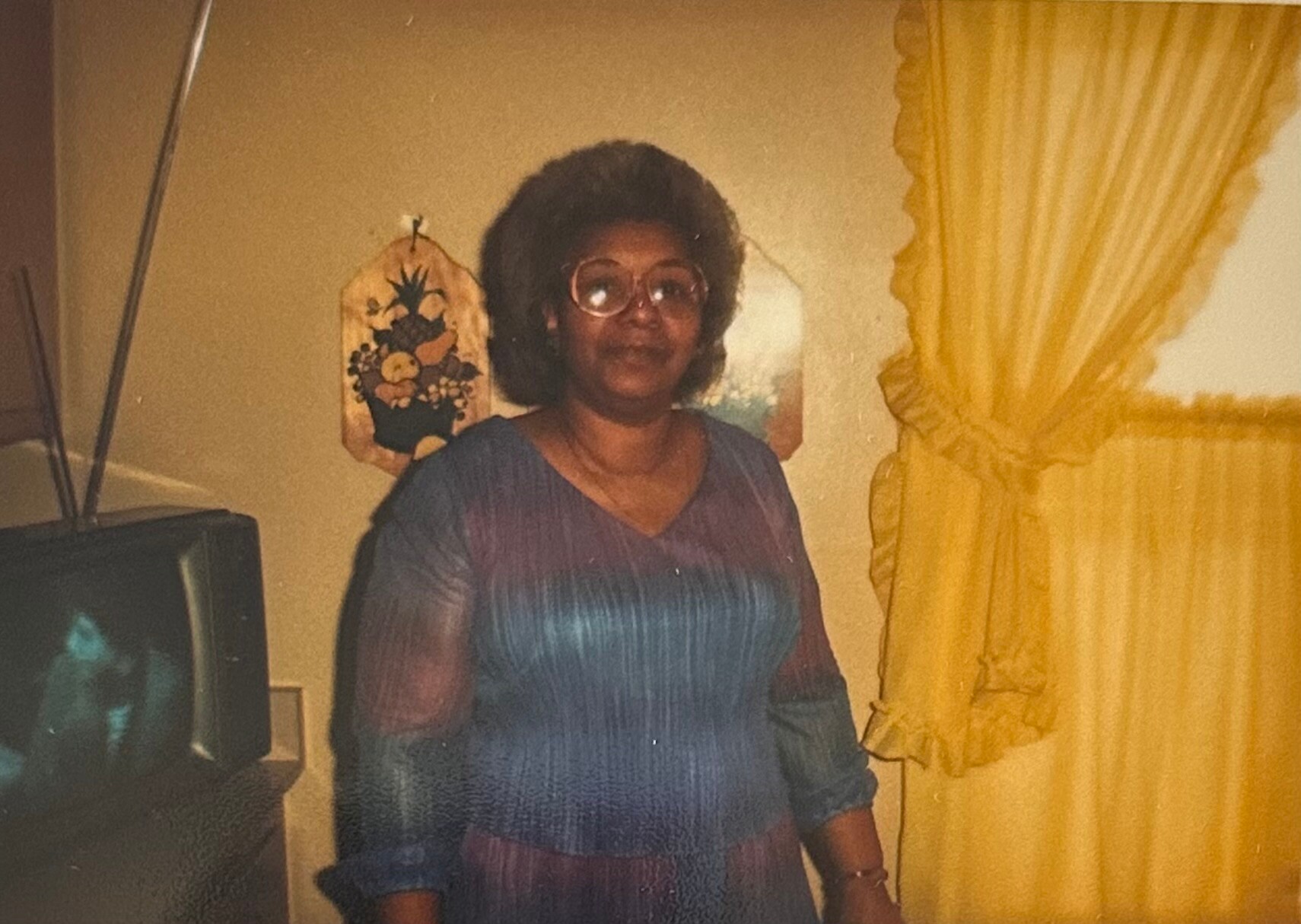 Woman with glasses in a blue dress standing indoors near yellow curtains and a vintage TV in the corner.