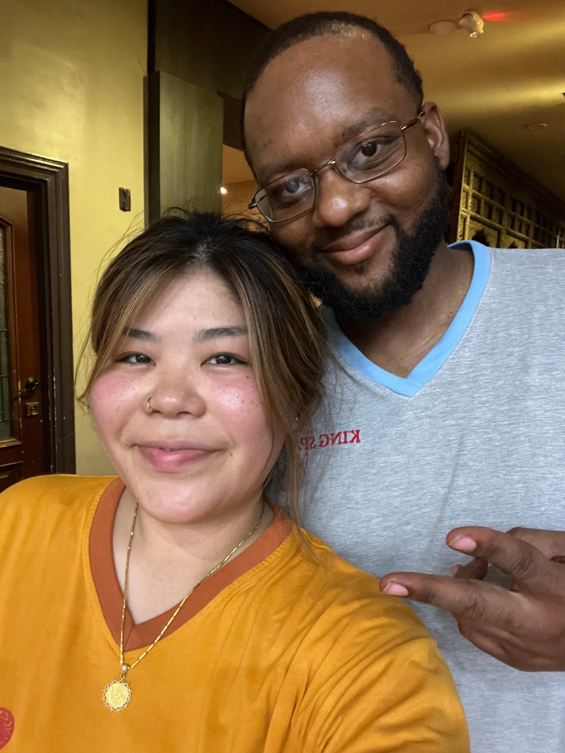 Two friends posing for a selfie indoors: woman in an orange shirt in the foreground and a bearded man with glasses behind her, flashing a peace sign.