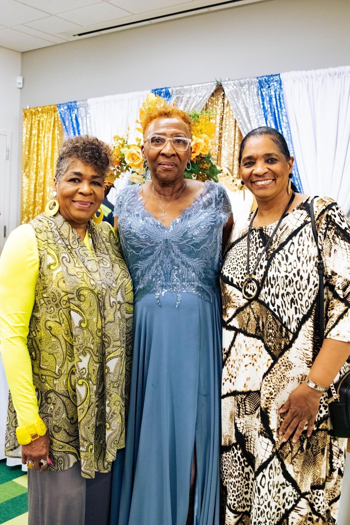 Three smiling women pose together at a celebration, with gold, blue, white sequin backdrops and a floral arrangement in the center behind them.